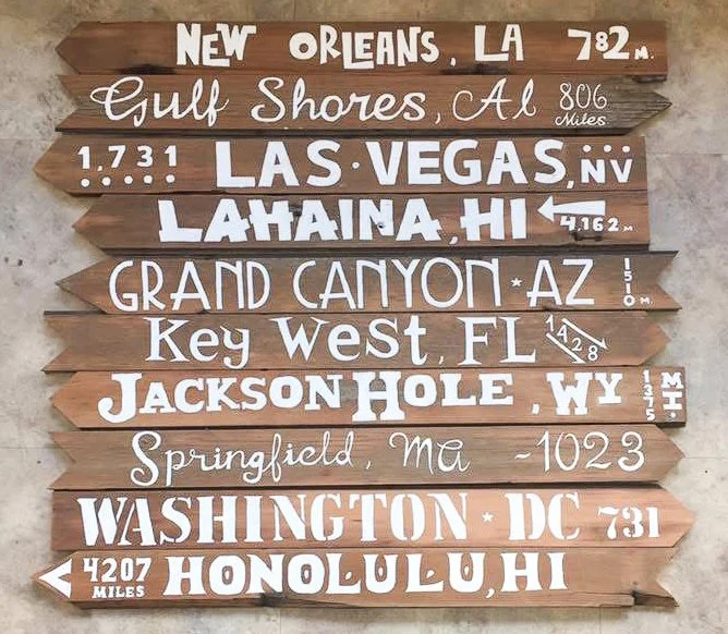 Decorative wooden signboard with travel destinations and distances in white lettering, including New Orleans, LA; Gulf Shores, AL; Las Vegas, NV; Lahaina, HI; Grand Canyon, AZ; Key West, FL; Jackson Hole, WY; Springfield, MA; Washington, DC; Honolulu, HI.