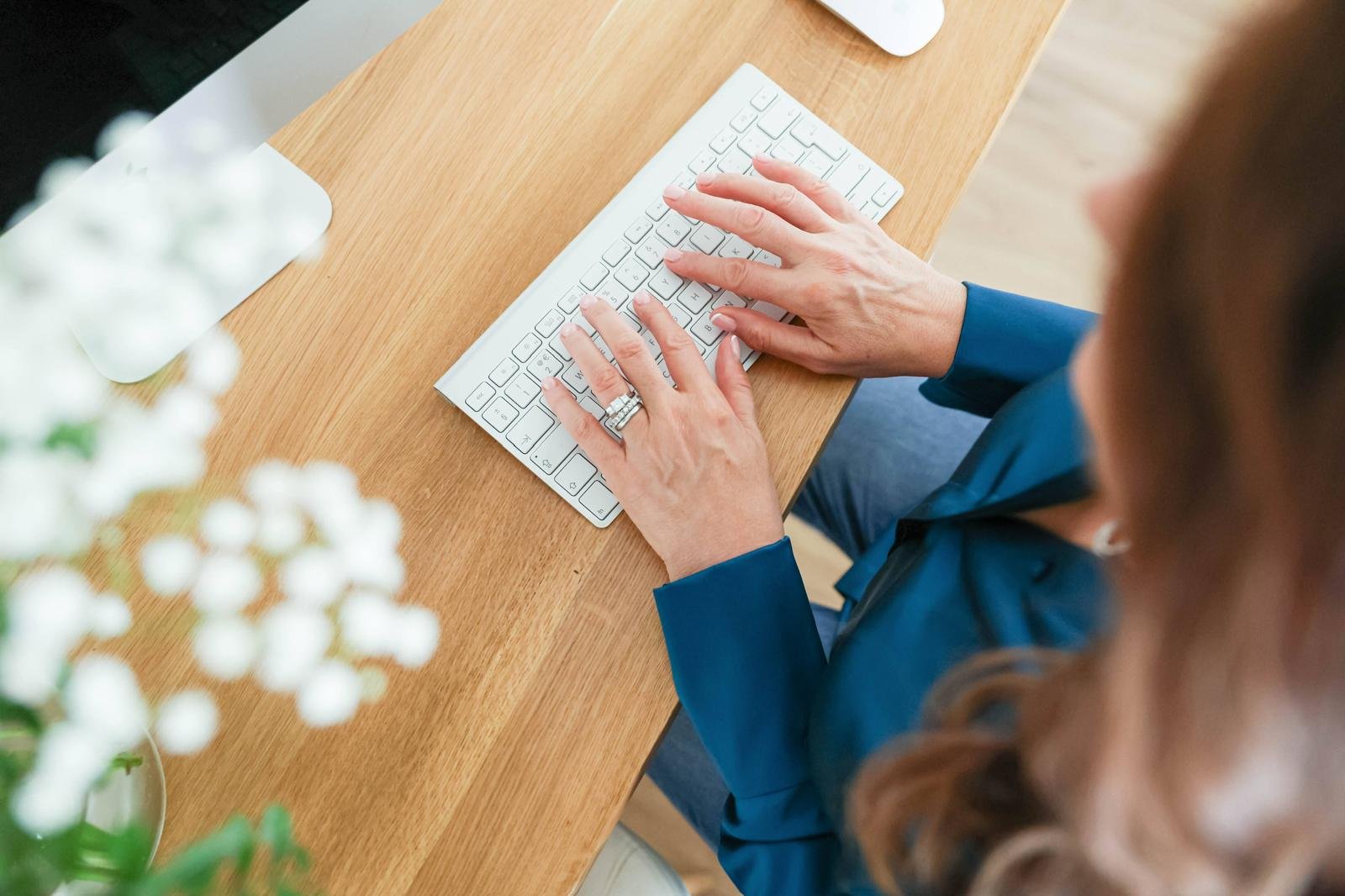 Person typing on a white wireless keyboard placed on a wooden desk, with a mouse nearby and white flowers in the foreground.