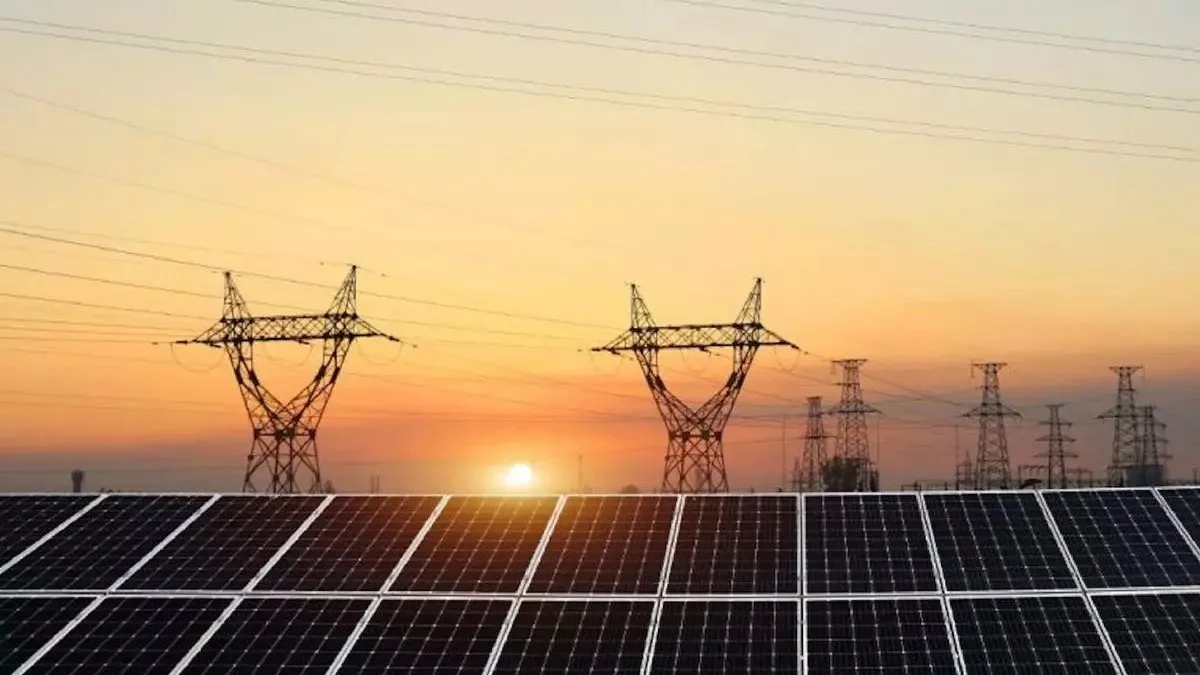 Solar panels in the foreground with power lines and transmission towers at sunset in the background.