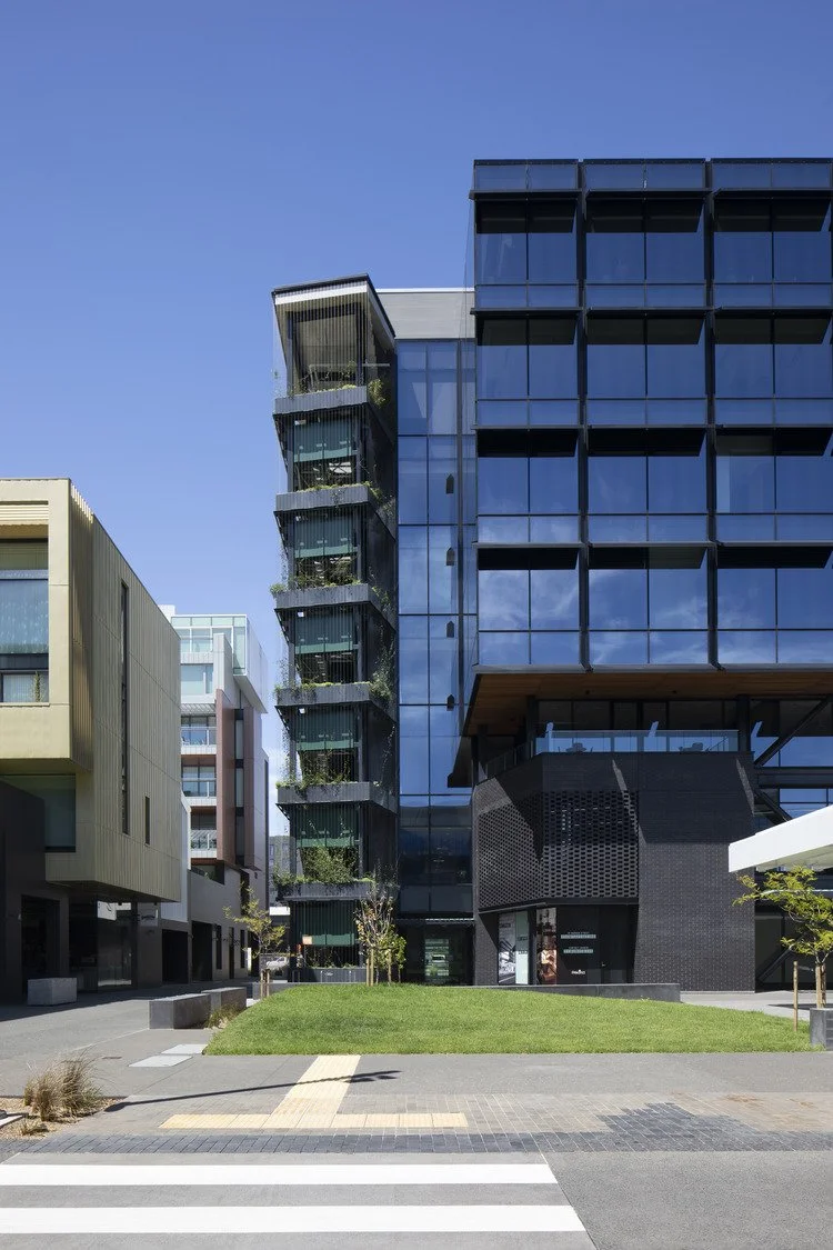 Modern multi-story office building with reflective glass windows and a rooftop terrace, situated on a city street with a small grassy area and pedestrian crosswalk.