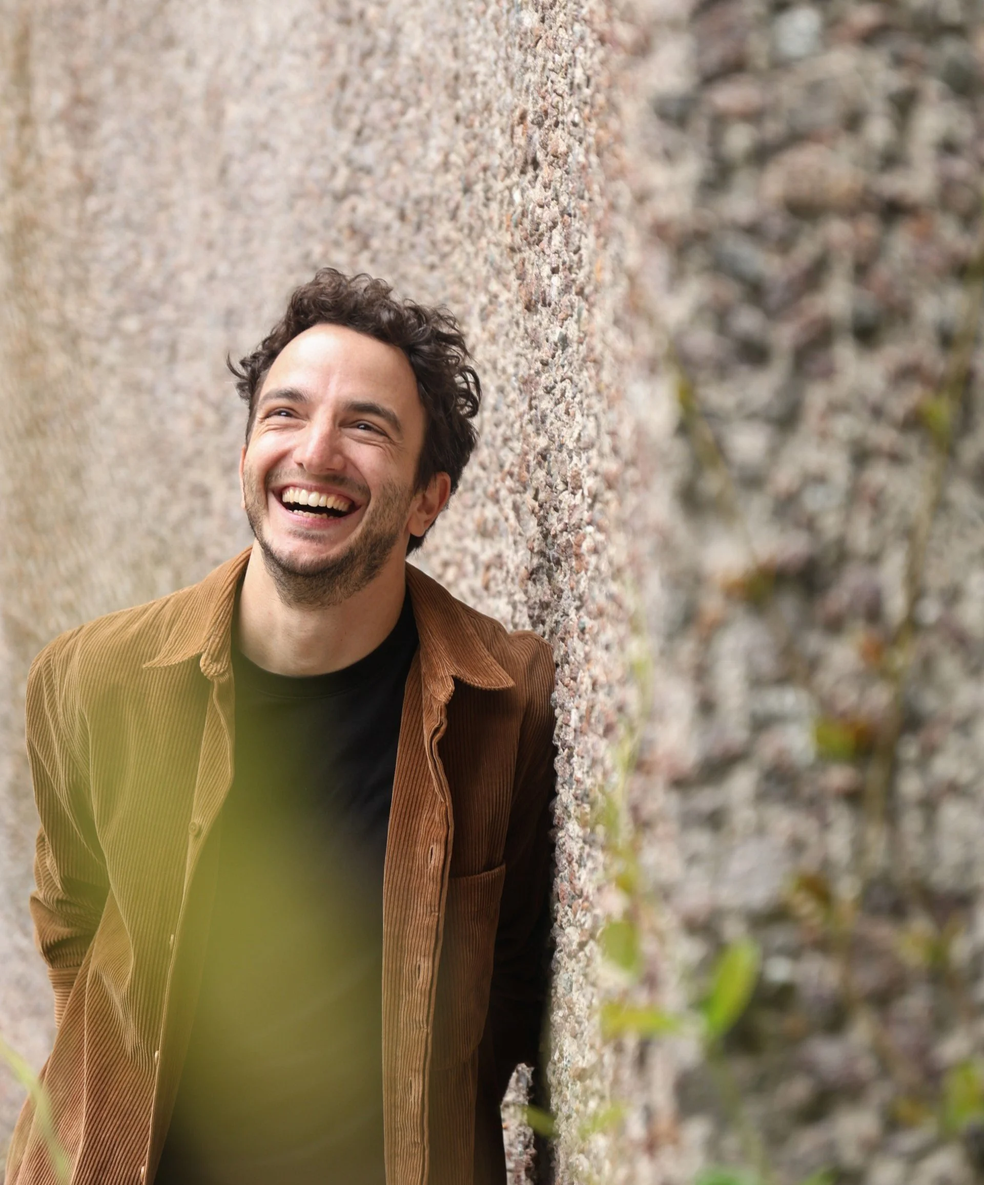 Ragazzo sorridente che si appoggia a un muro di pietra con sfondo naturale.