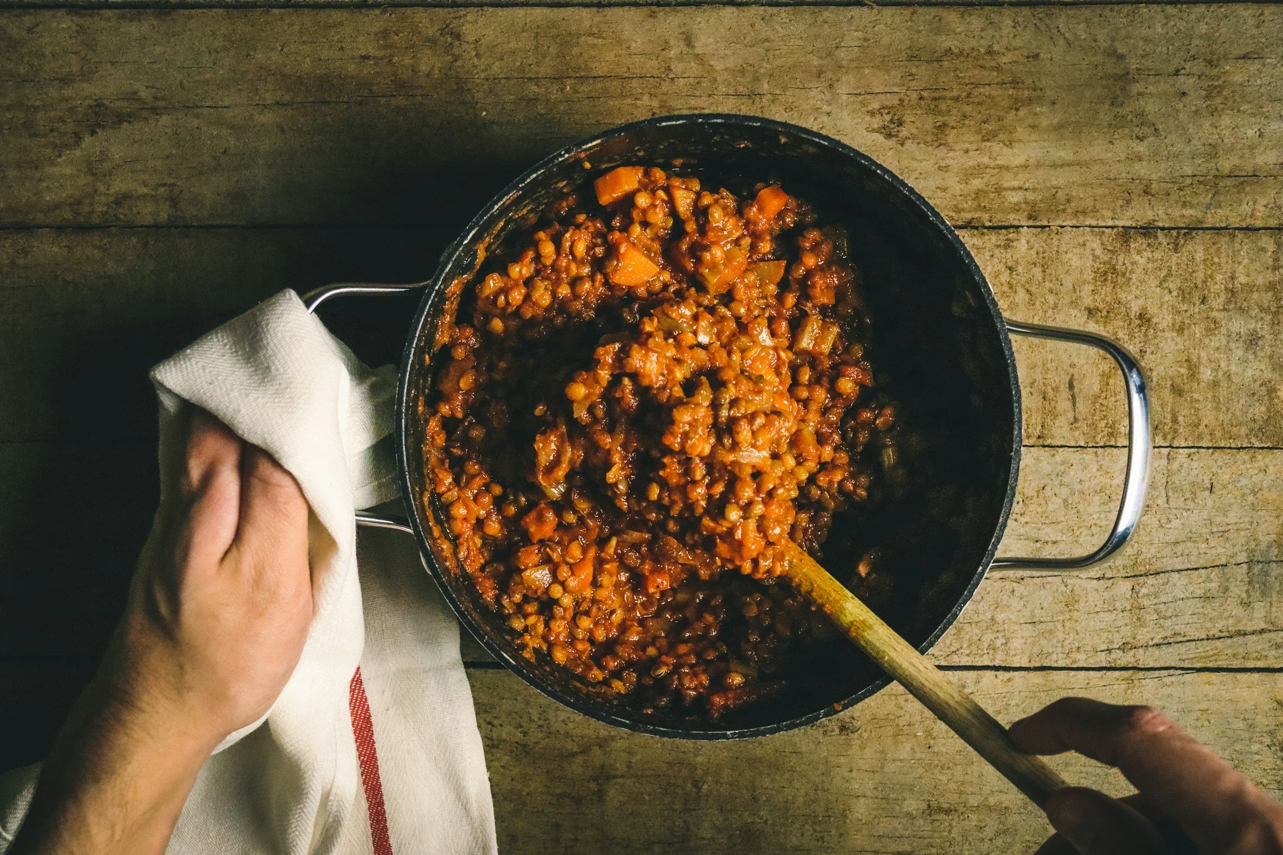 a hand and wooden spoon mixing a meal of lentils in a pan