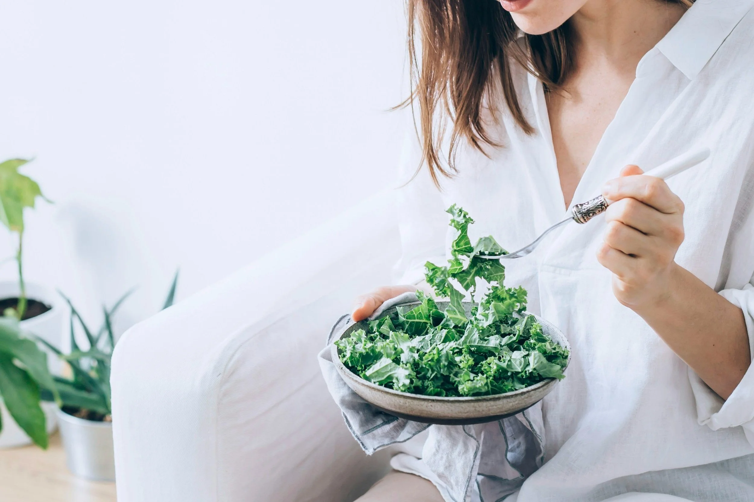 A woman is sitting on a white couch, holding a bowl of mixed leafy greens, and eating a salad with a fork.