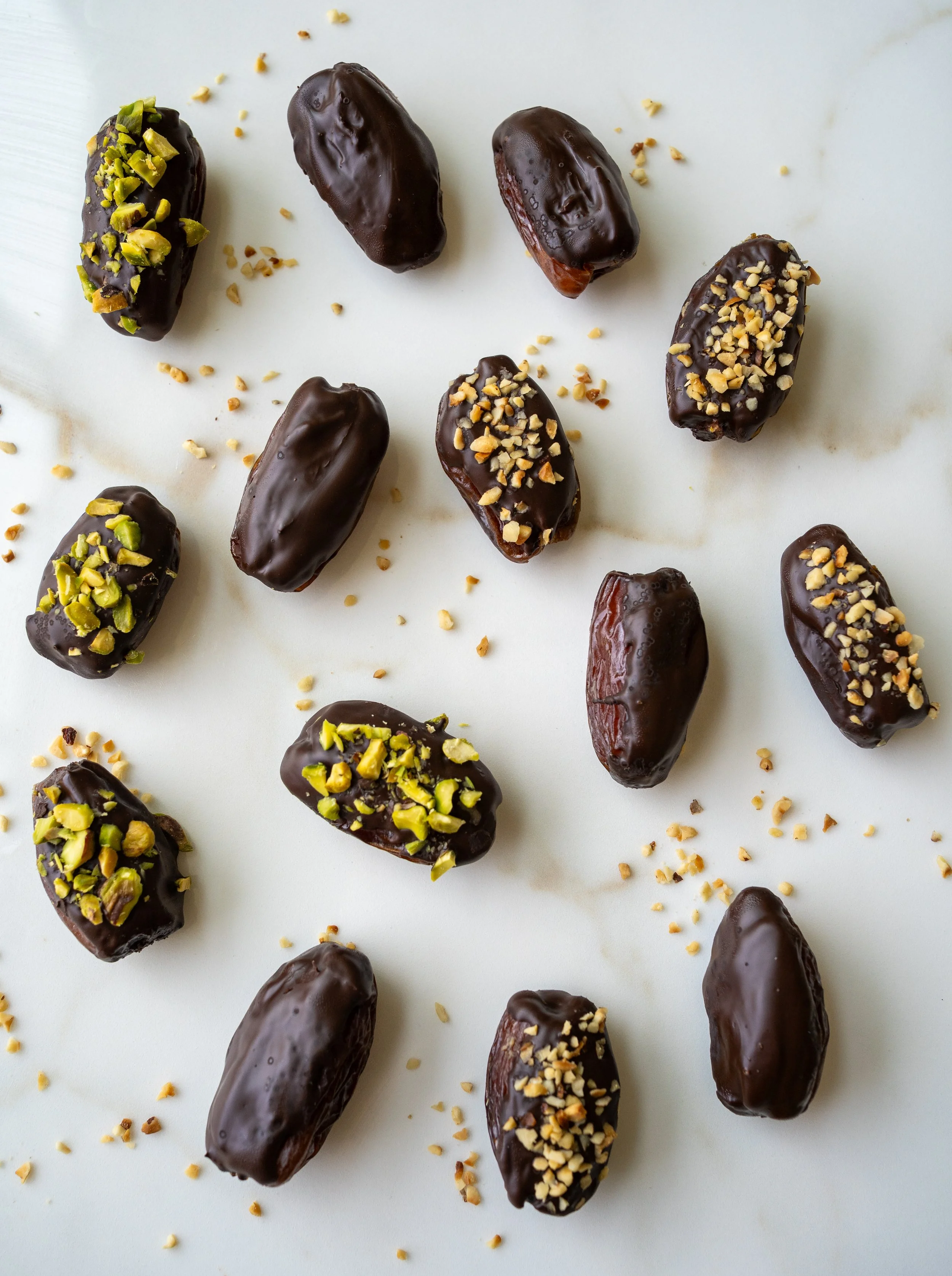 Chocolate covered stuffed dates sprinkled with nuts on a white marble surface