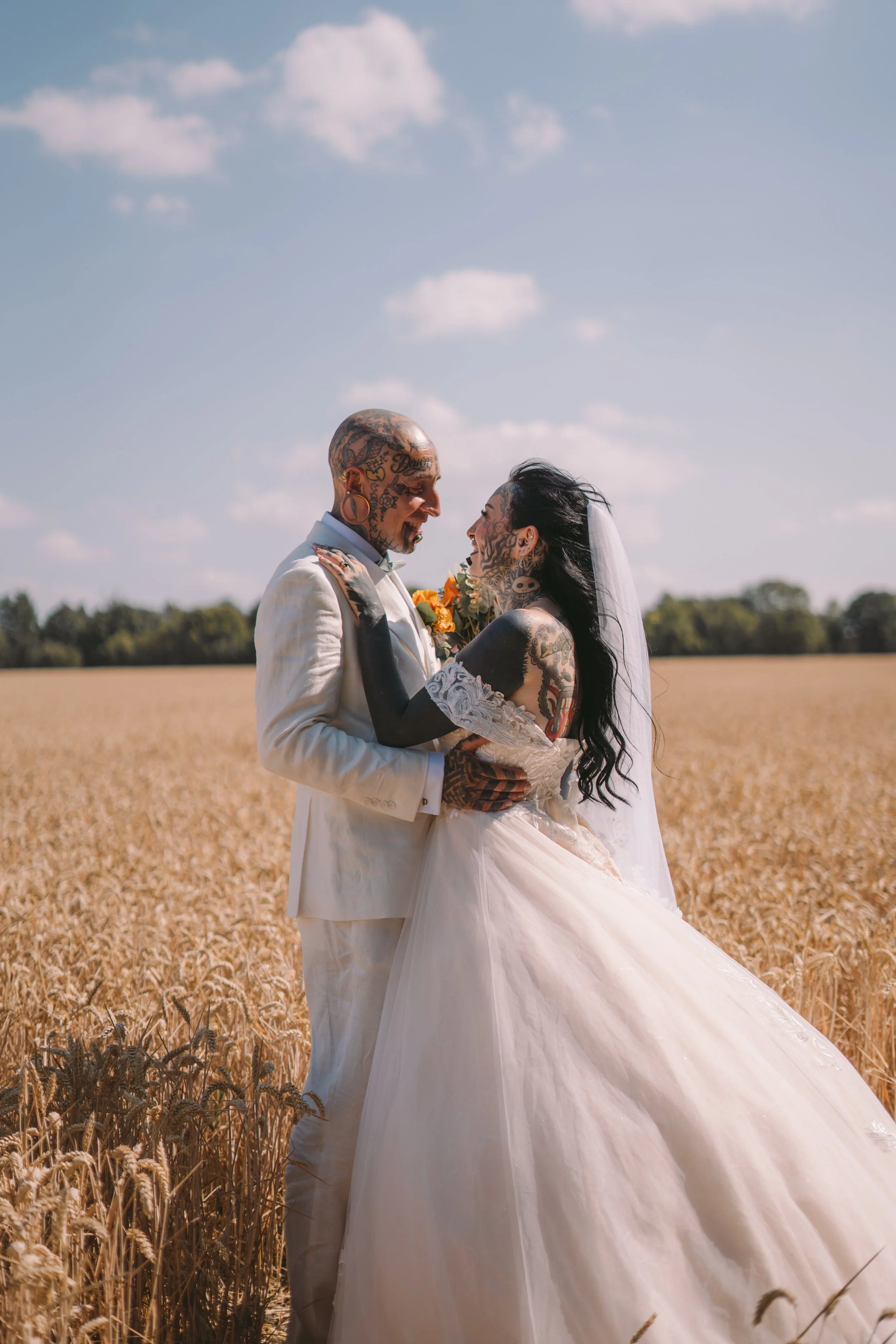 A newlywed couple, both heavily tattooed, standing close together in a golden wheat field under a partly cloudy blue sky, gazing into each other's eyes.