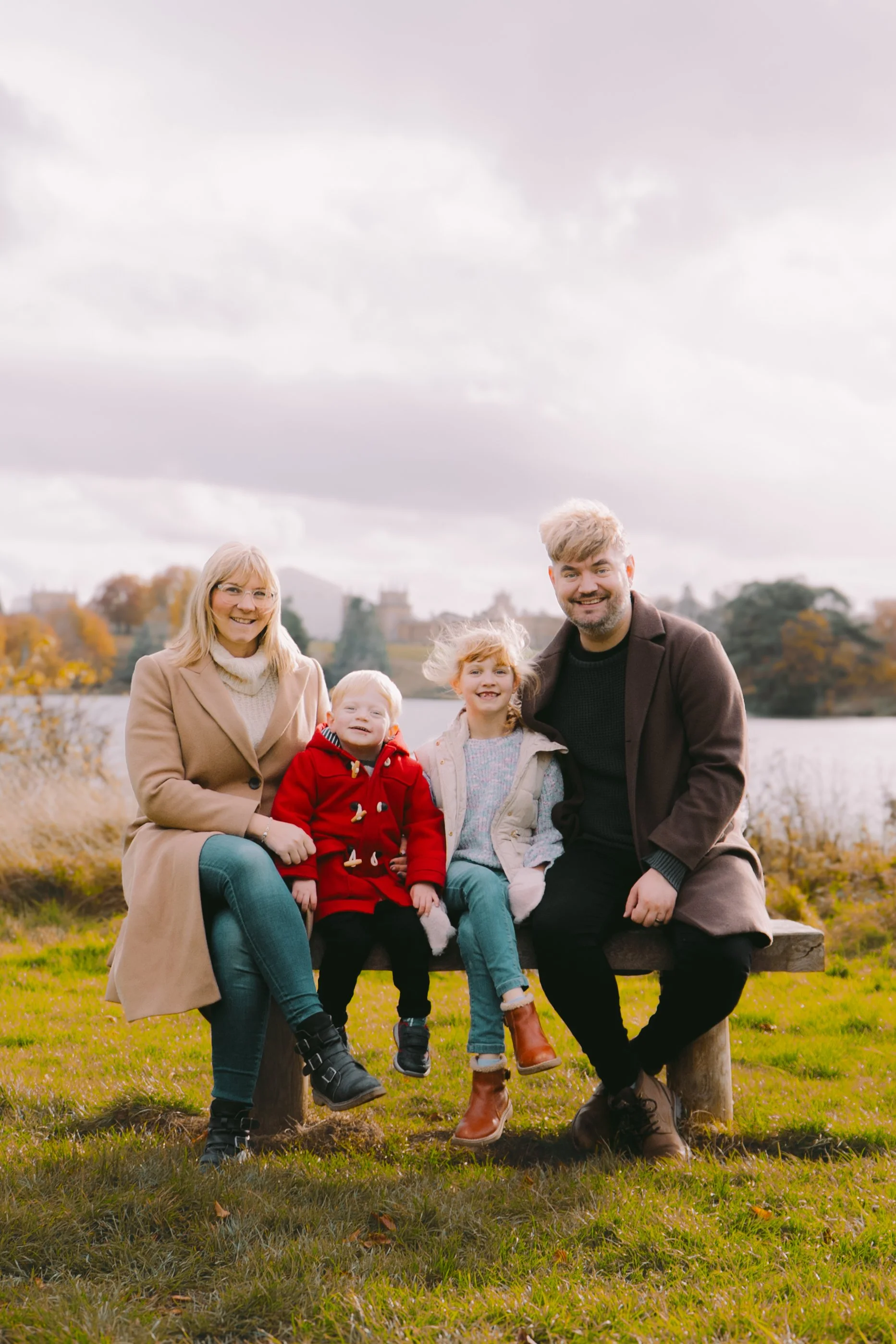 A family of five sitting outdoors on a bench near water with trees and a castle in the background, cloudy sky, autumn foliage, all smiling.