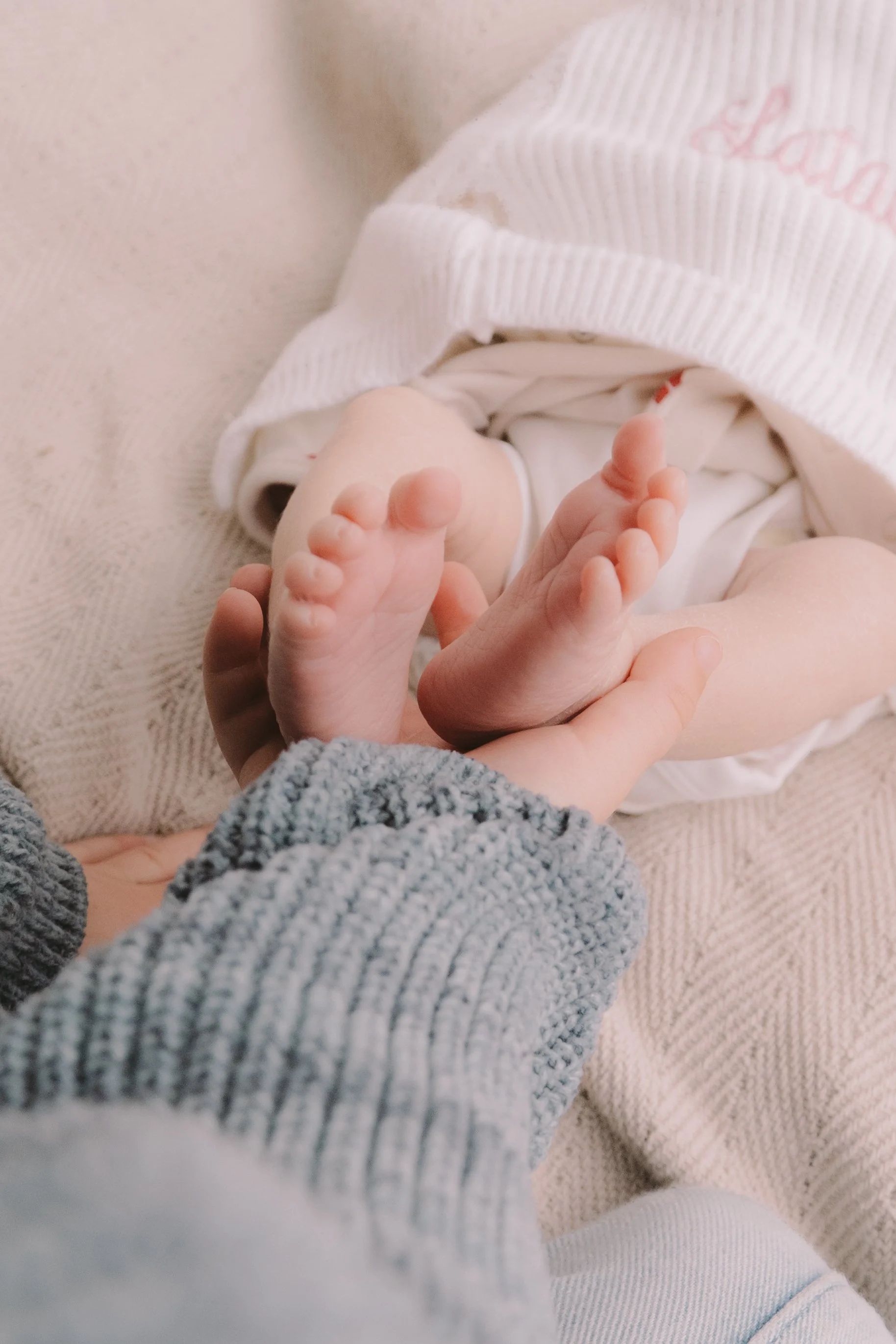 Close-up of a adult hand gently holding a baby's tiny feet, with the baby wearing a white outfit and sitting on a cozy surface.