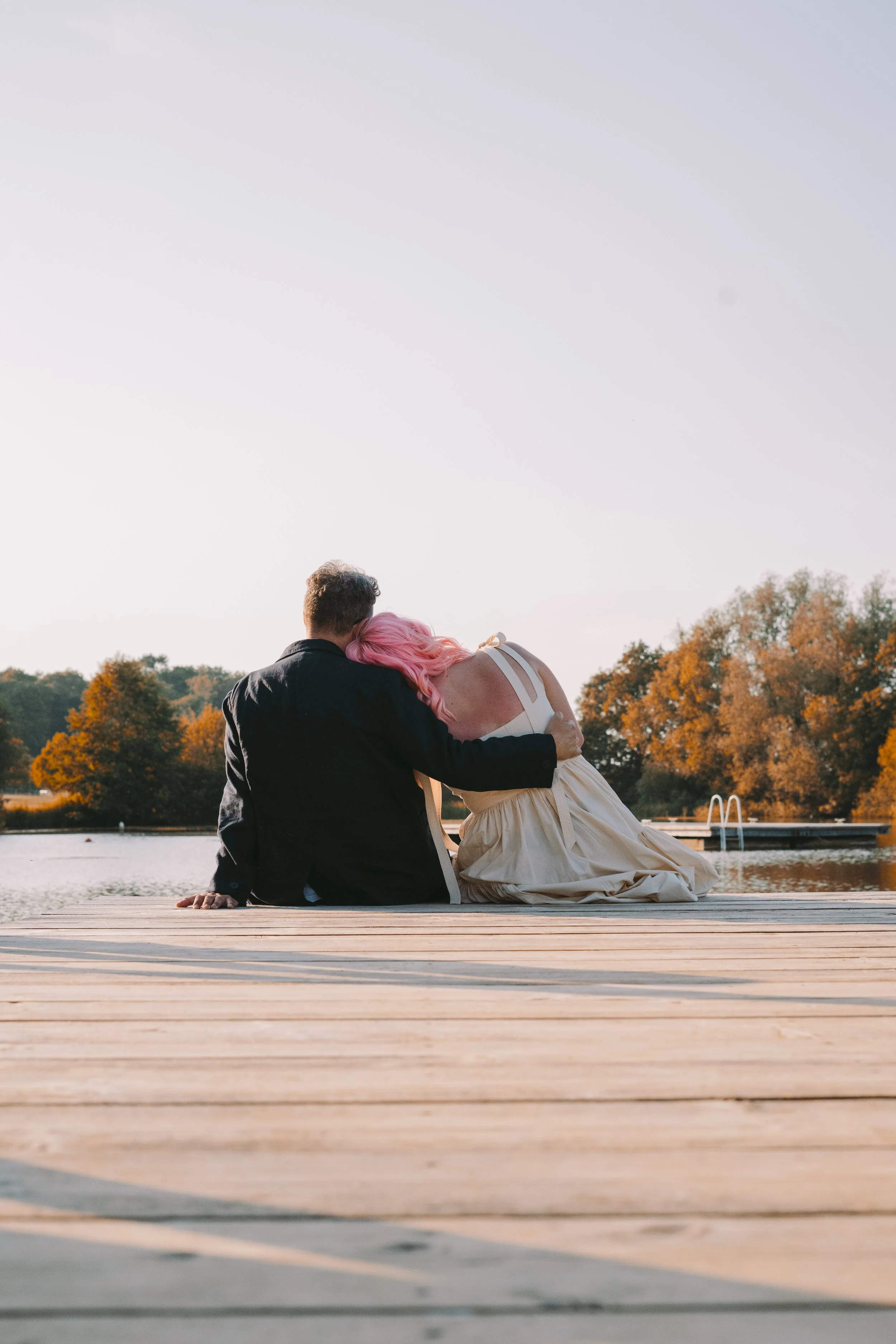 A couple sitting by a lake on a dock, with the man wearing a dark jacket and the woman with pink hair in a white dress, embraced with the back to the camera, surrounded by fall foliage.