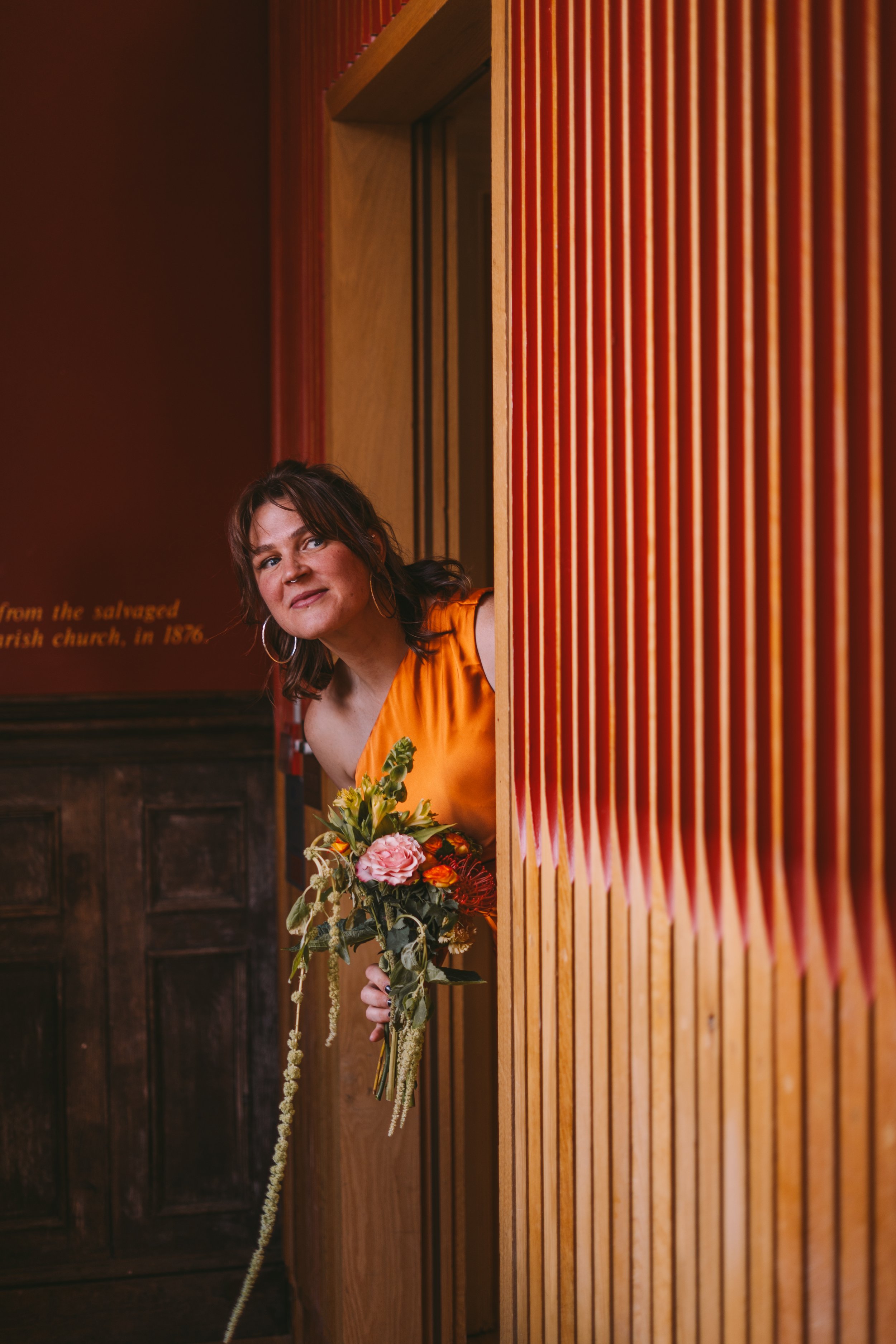 A woman with dark hair, wearing a yellow dress, holding a colorful floral bouquet, peering around a wooden partition in an indoor setting with dark wooden walls.