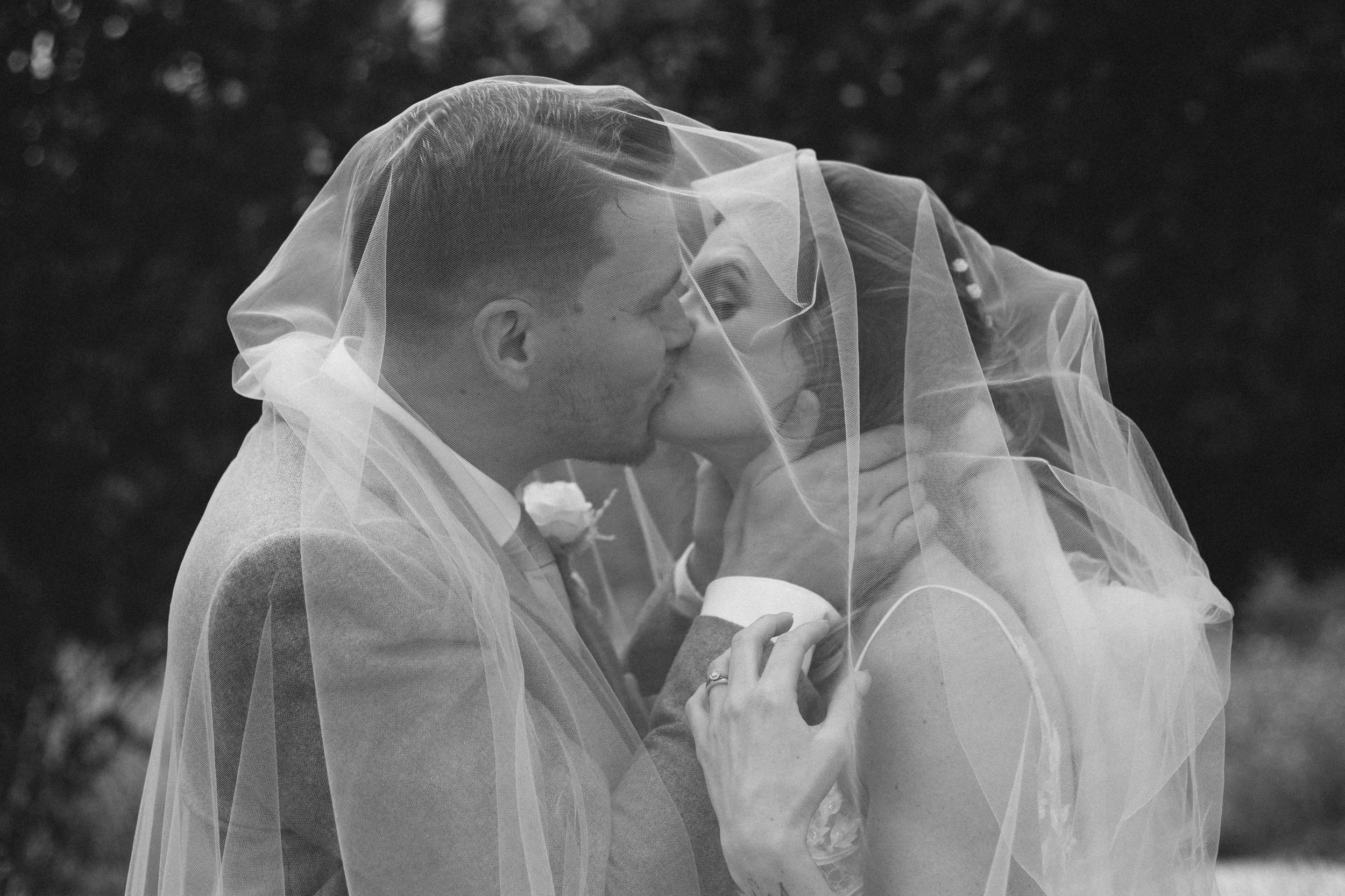 A black and white photo of a couple, with the groom dressed in a suit and the bride wearing a veil, sharing a kiss outdoors.