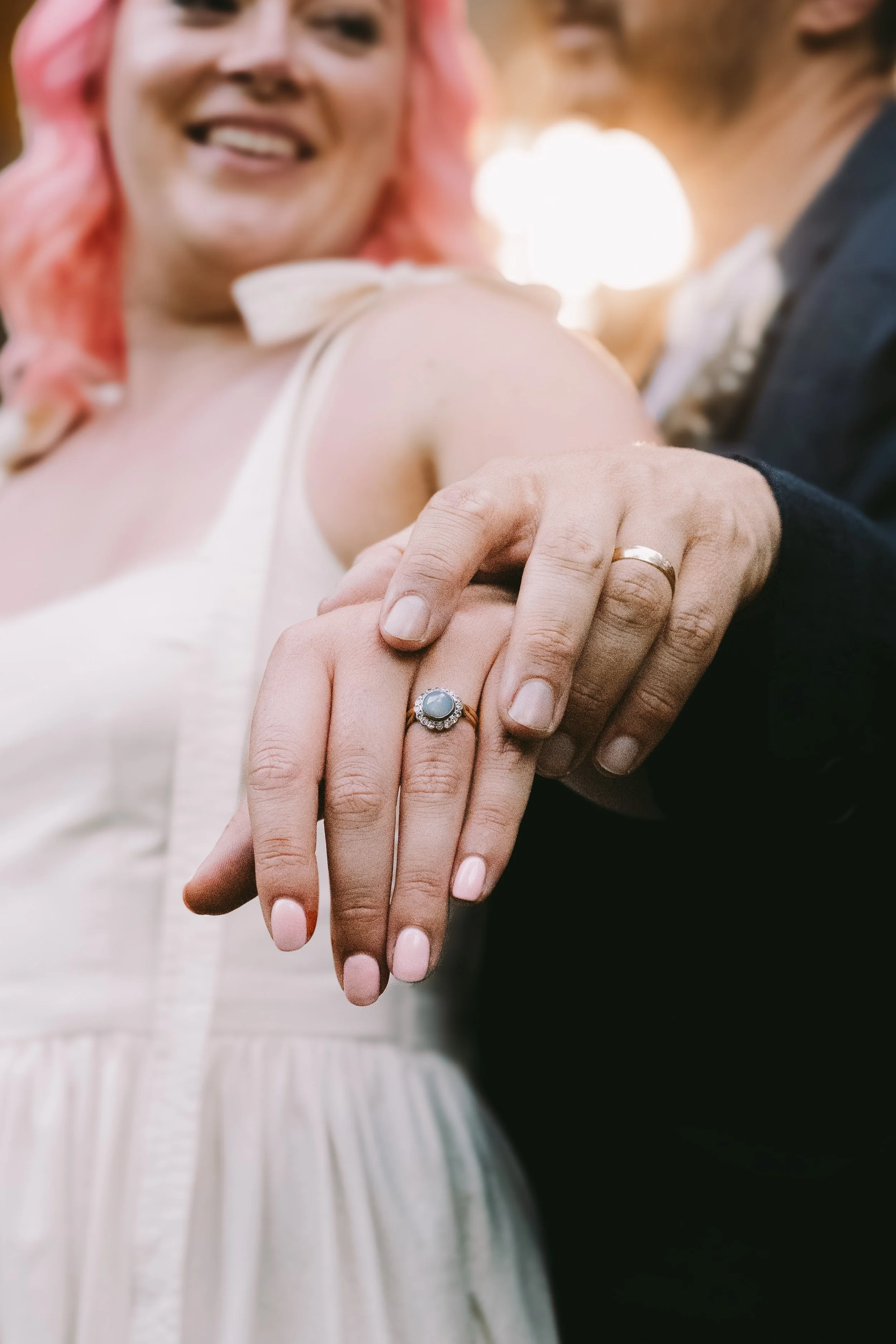 A couple holding hands, showing wedding rings, with a woman smiling in the background.