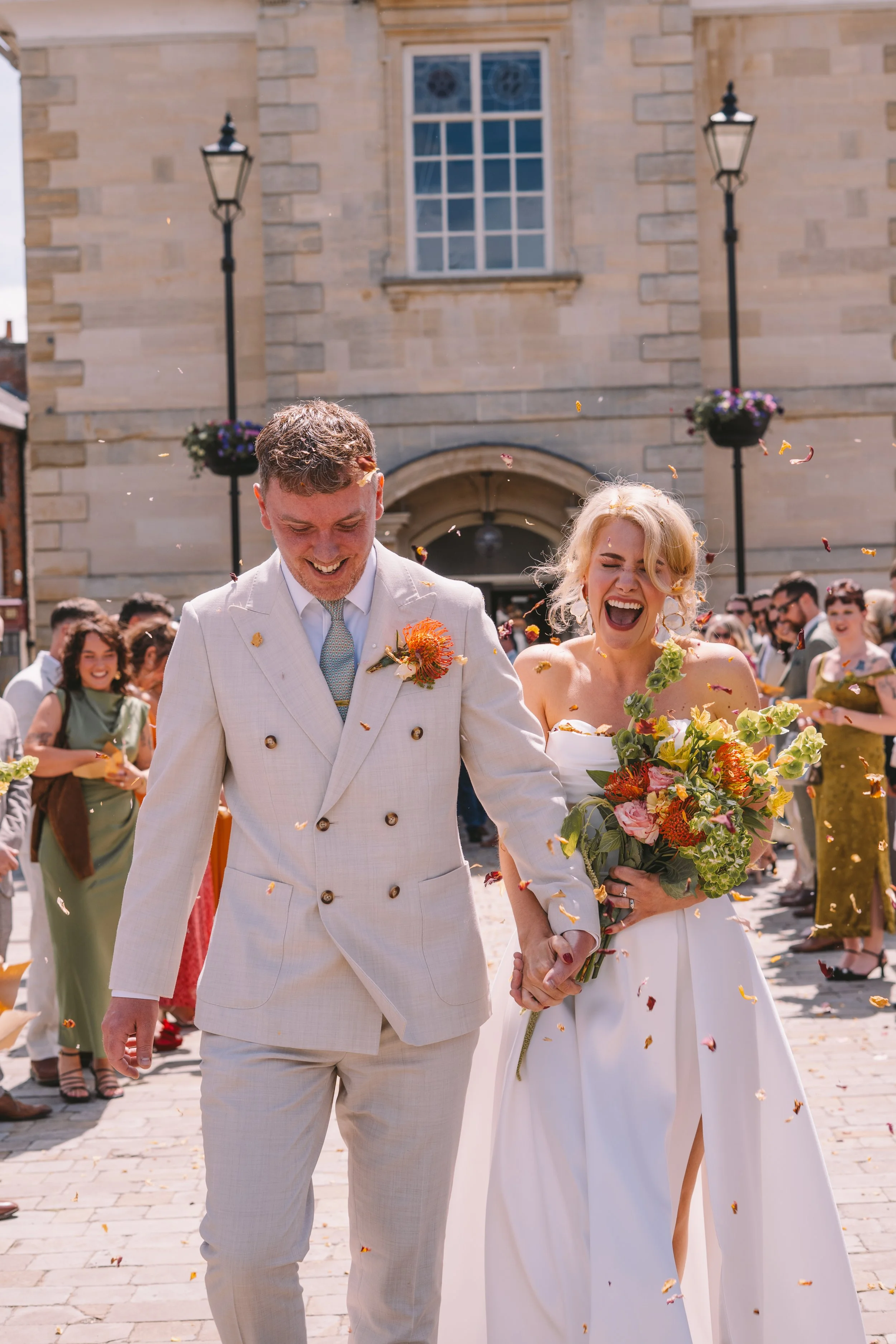 A newly married couple holding hands and smiling, surrounded by friends, tossing flower petals in a street in front of a historic stone building with large windows and two lamp posts with flower baskets.