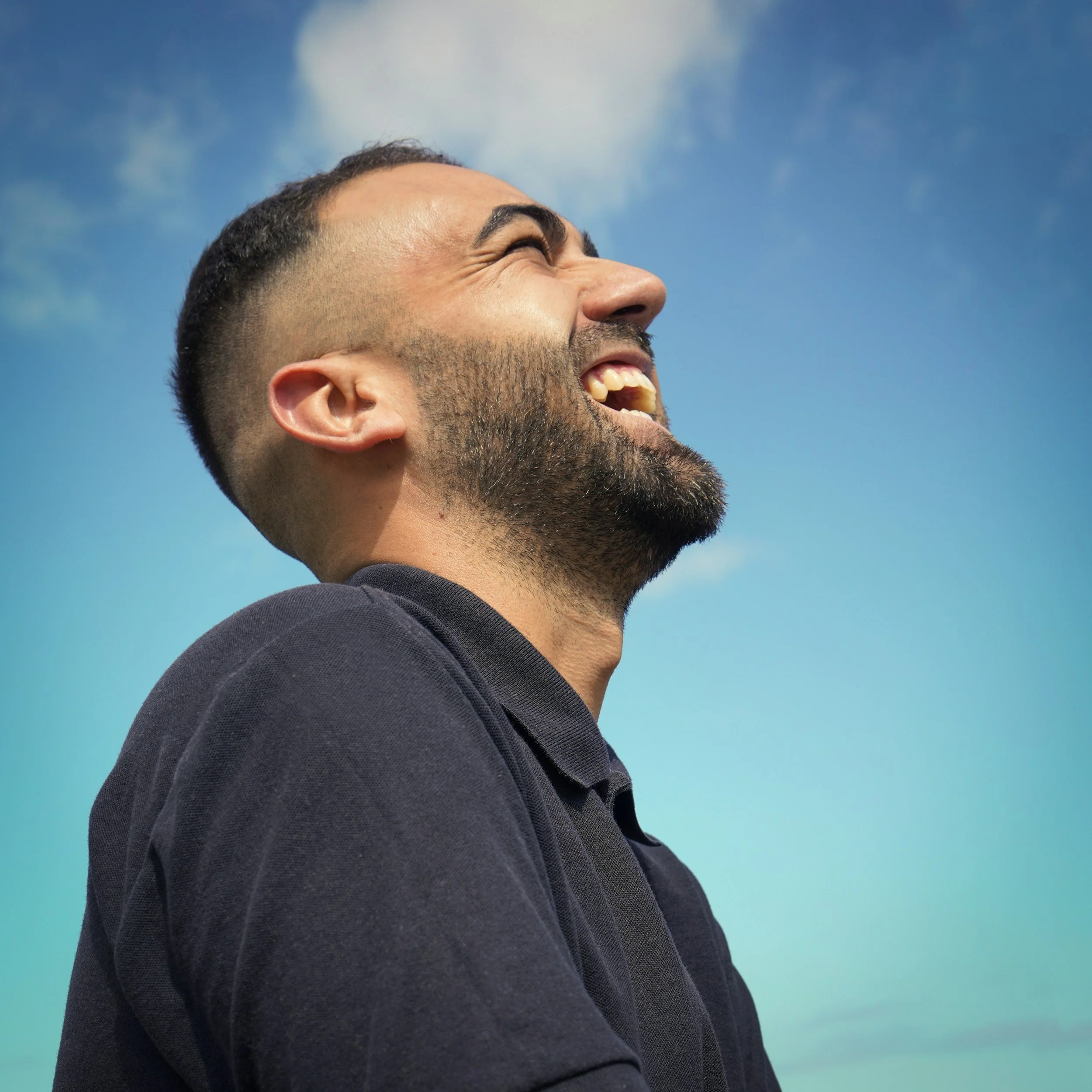 A man with a beard and short hair, smiling and looking up, against a blue sky with some clouds.