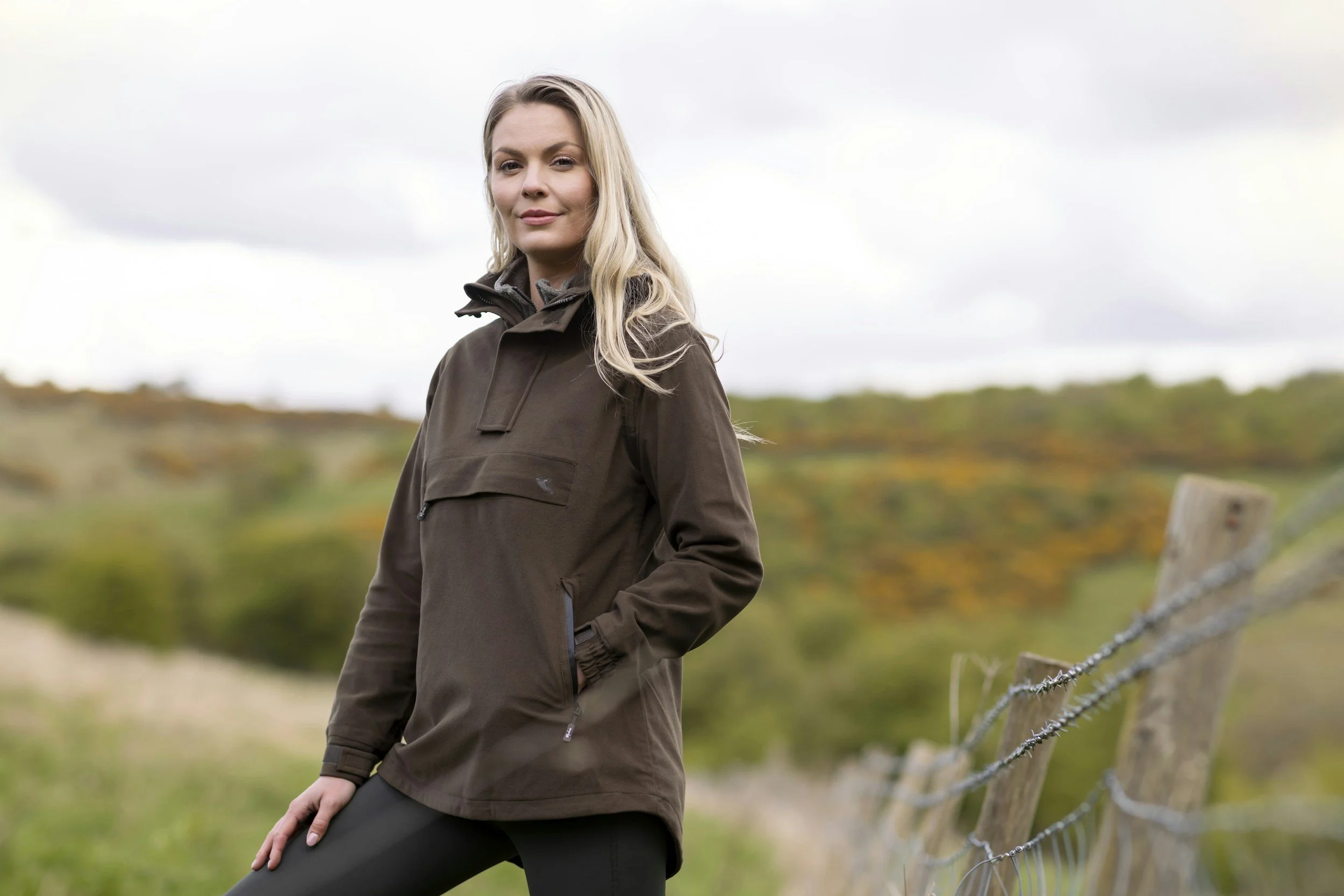 A woman standing outdoors near a wooden fence with wire, wearing a brown jacket and black pants, with a blurred background of trees and hills.