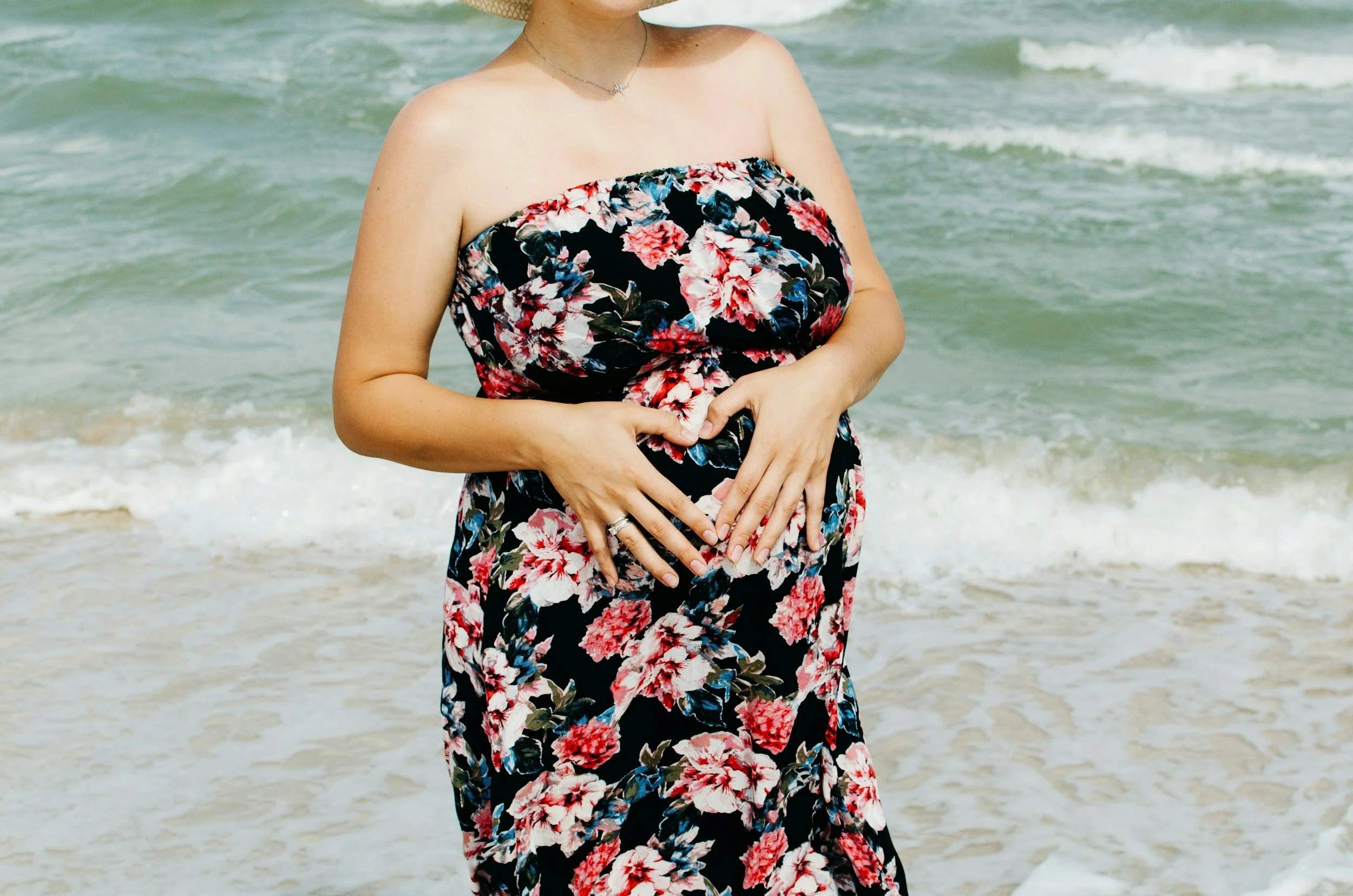 A woman on the beach wearing a strapless floral dress, with hands forming a heart shape over her abdomen, standing near the ocean waves.