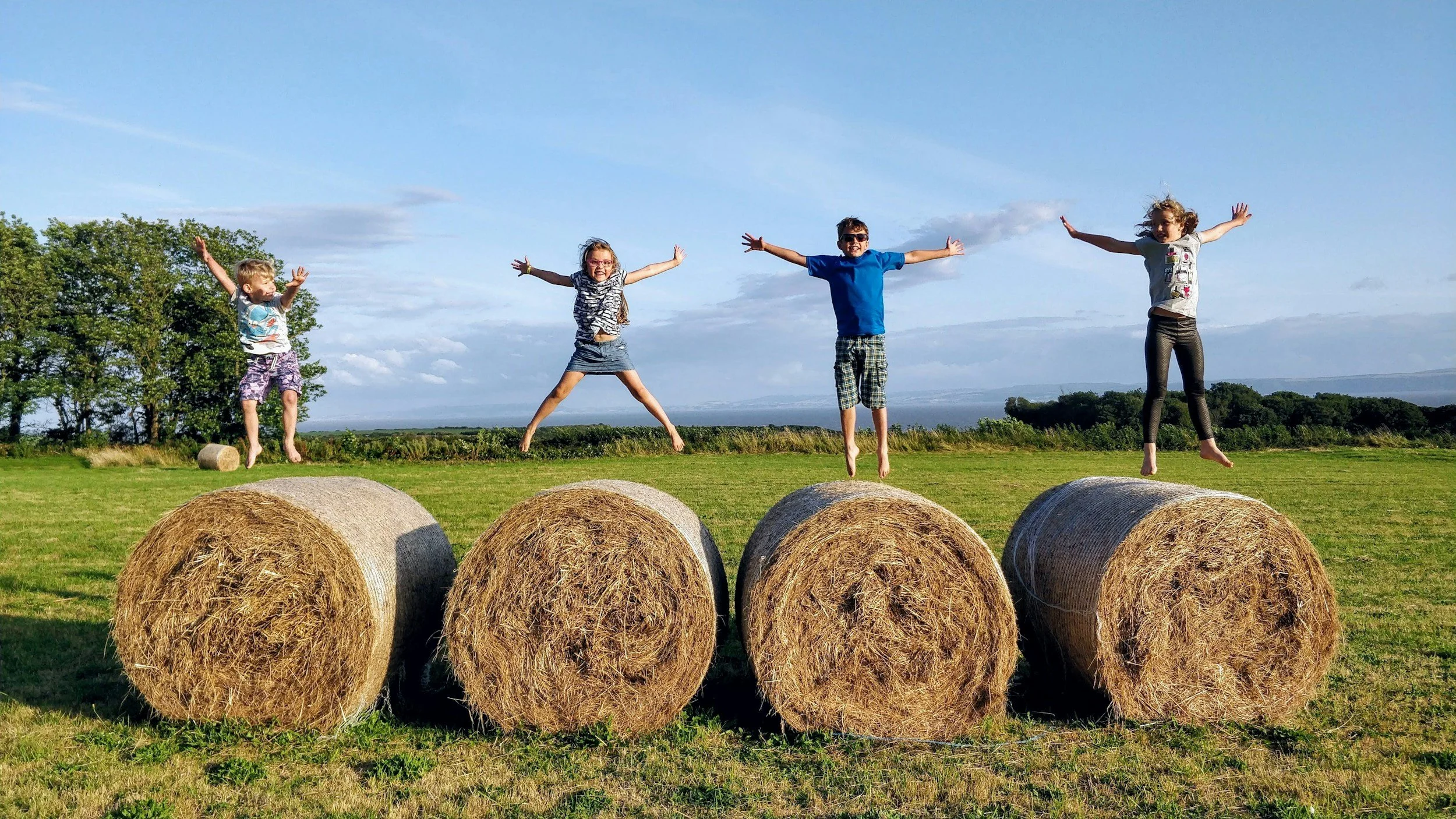 Four children jumping on hay bales in a grassy field under a blue sky with clouds.