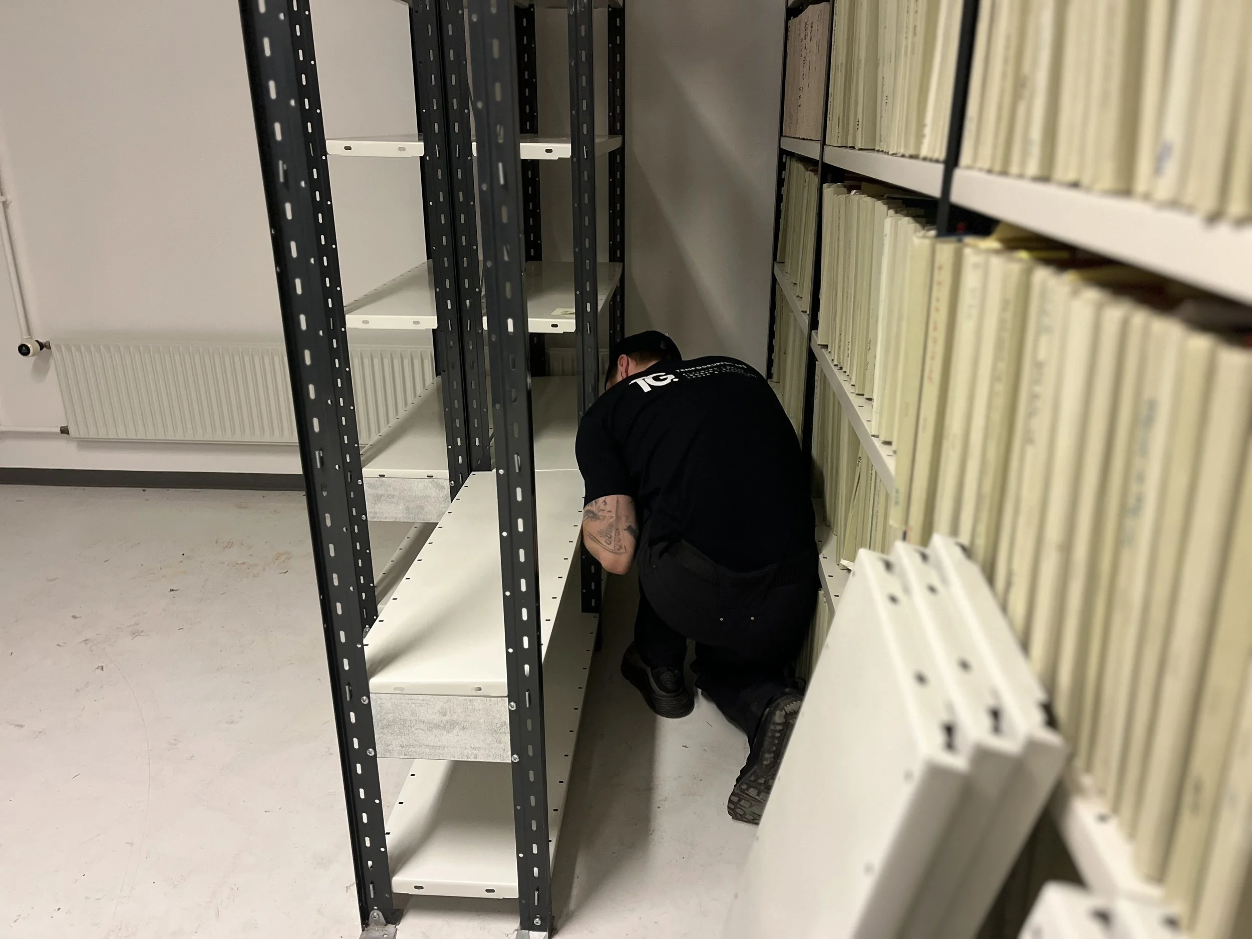 En person er på knæ i et bibliotek, opdager eller arbejder med bøger eller arkiver, placeret foran Tomme hylder.