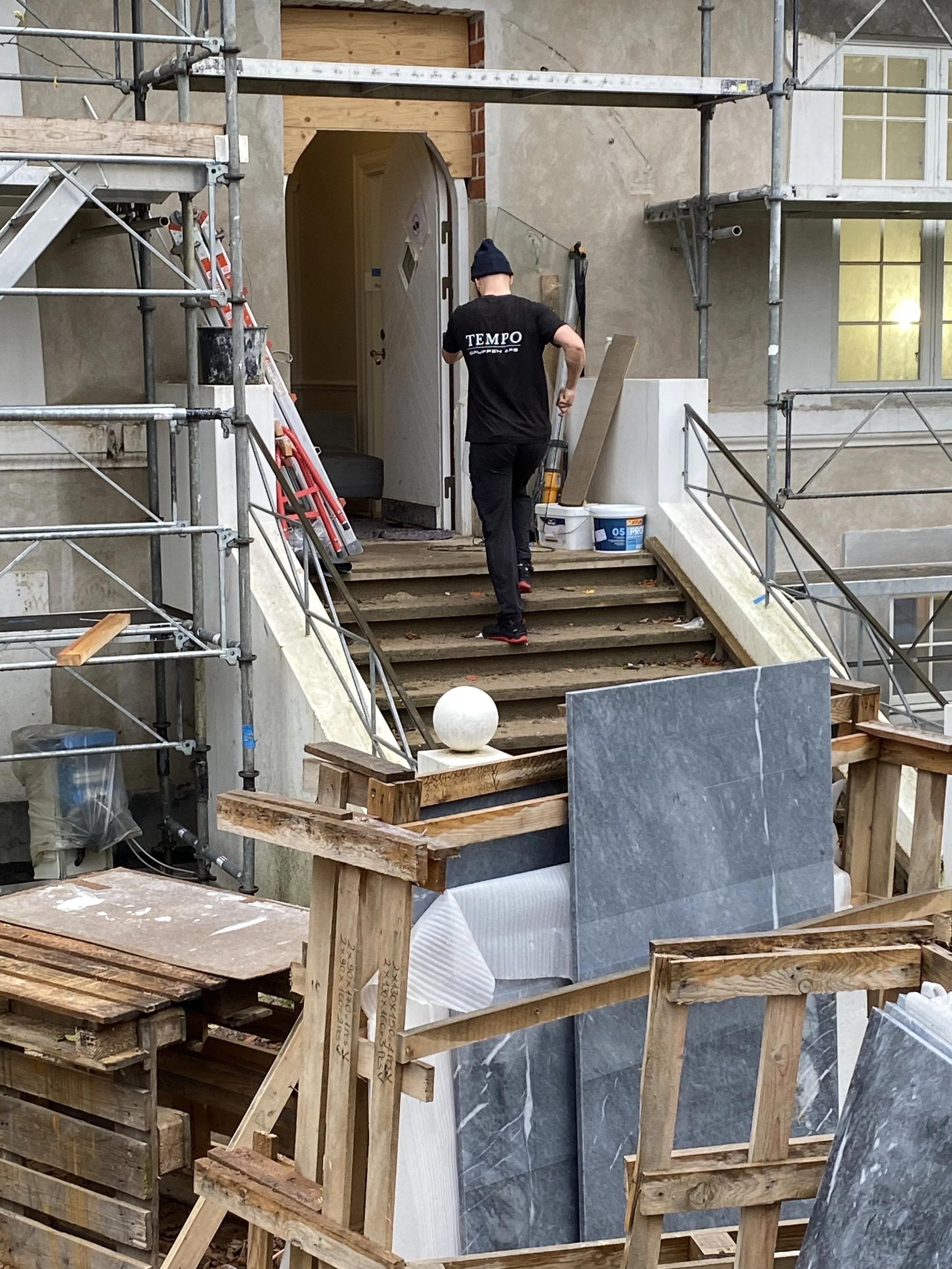 En person går op ad udendørs trappe under renovering af en bygning, omgivet af byggeudstyr og stillads.