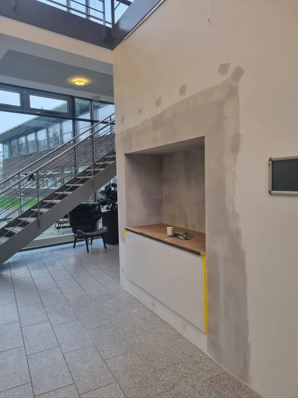 Indendørs område med trappe, vinduer, en stol og en væg under konstruktion eller renovering.