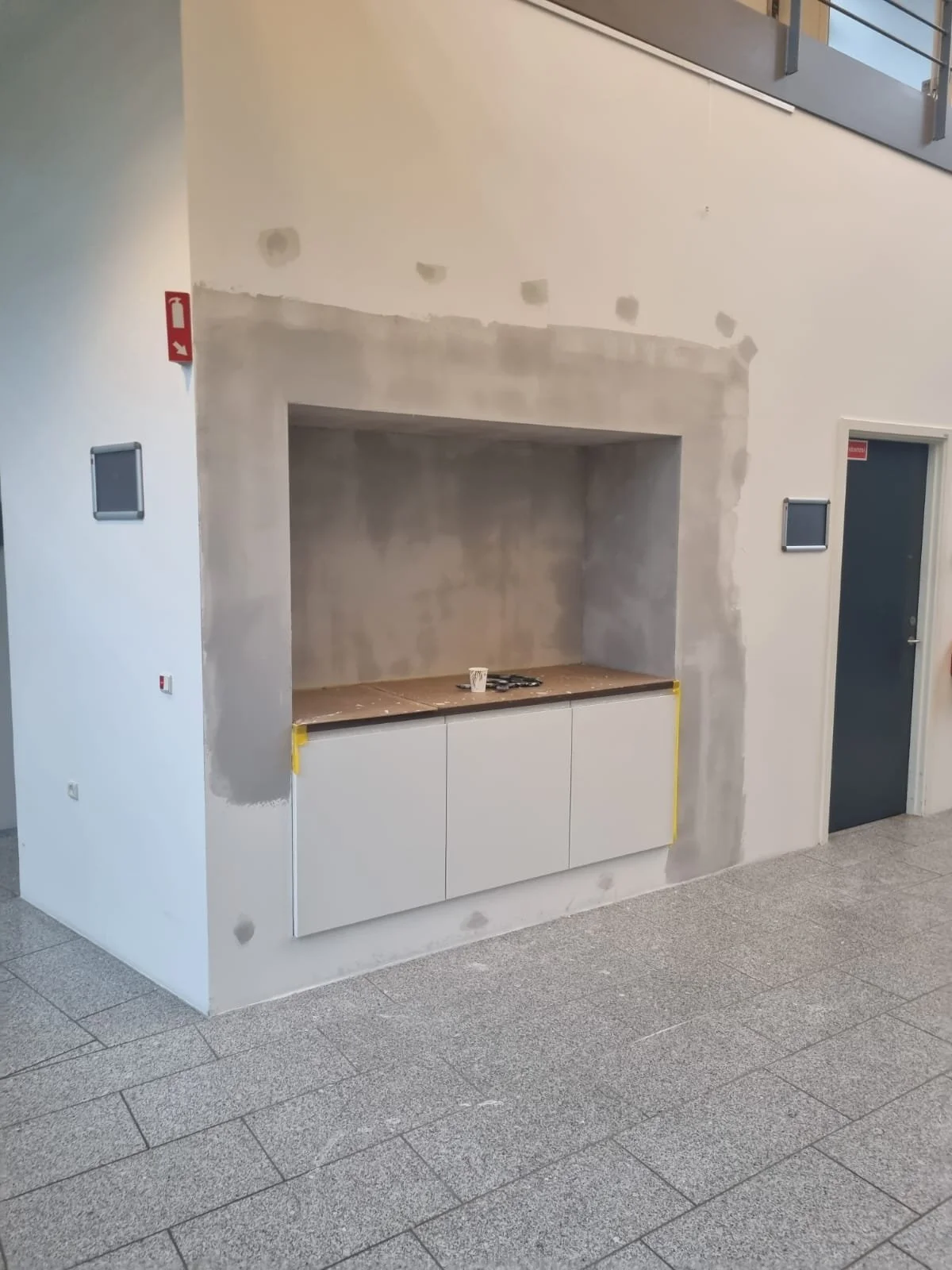 En moderne stue med en halvbygget kamin, væggen er under renovering med grå betonspor og en hvid væg. Der er to små sorte vægpaneler og et låst rumdør på højre side.