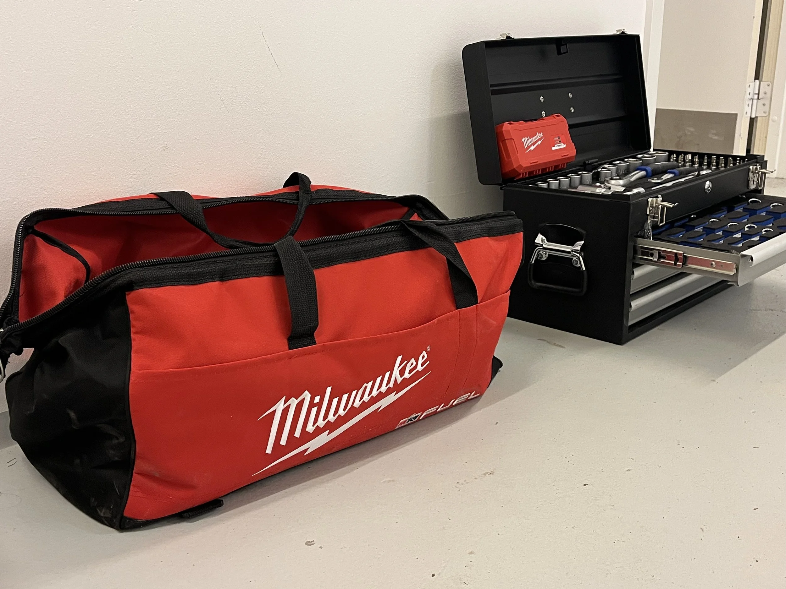 Rød værktøjstaske med Milwaukee logo ved siden af et sort værktøjskab med skruenøgler og skruer.