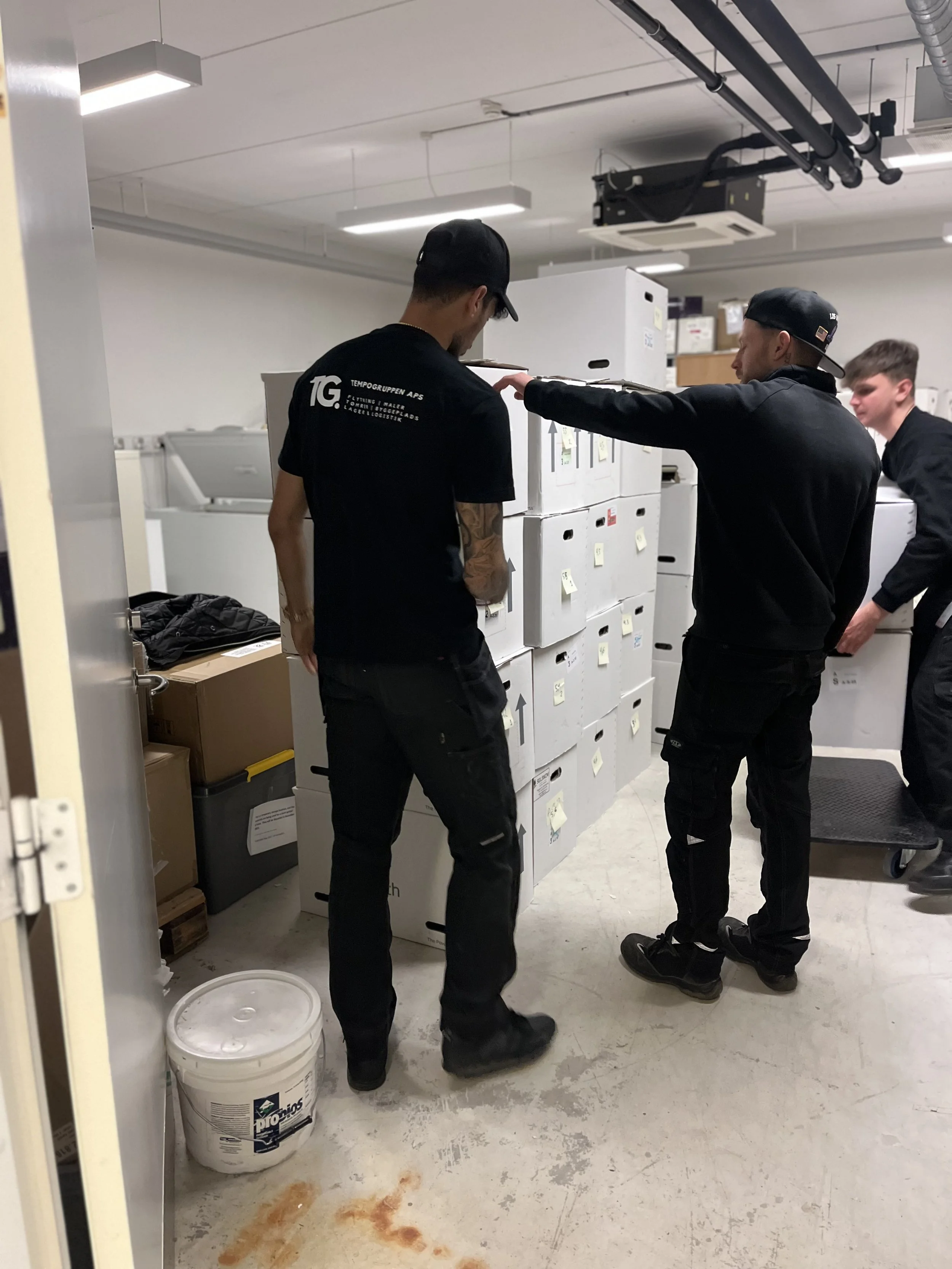 Tre personer står i en kælder eller et teknisk rum med hvide skabe og apparater. To af personer har caps, og de ser ud til at være i en samtale eller diskussion. Der er kasser, en bøtte og noget snavs på gulvet.