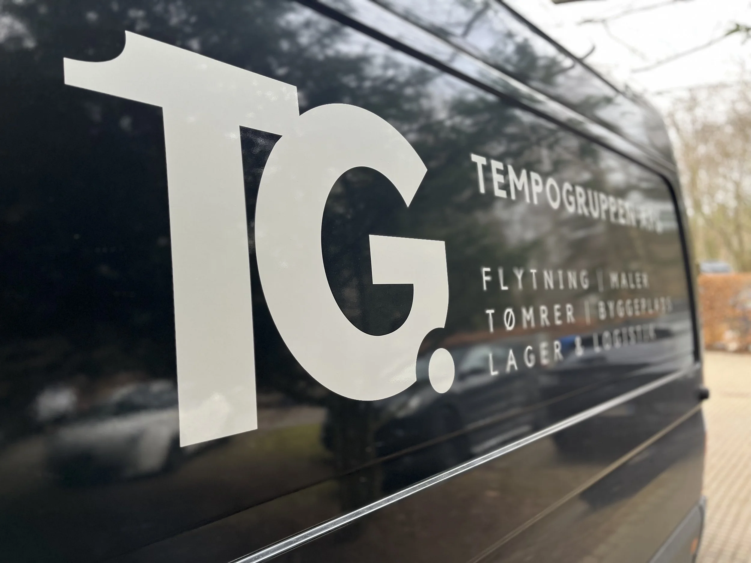 Sort servicevogn med hvide tekst, herunder logo'et for TG, og forskellige tjenester listet, såsom flytning, tømrer, bygger og lager.