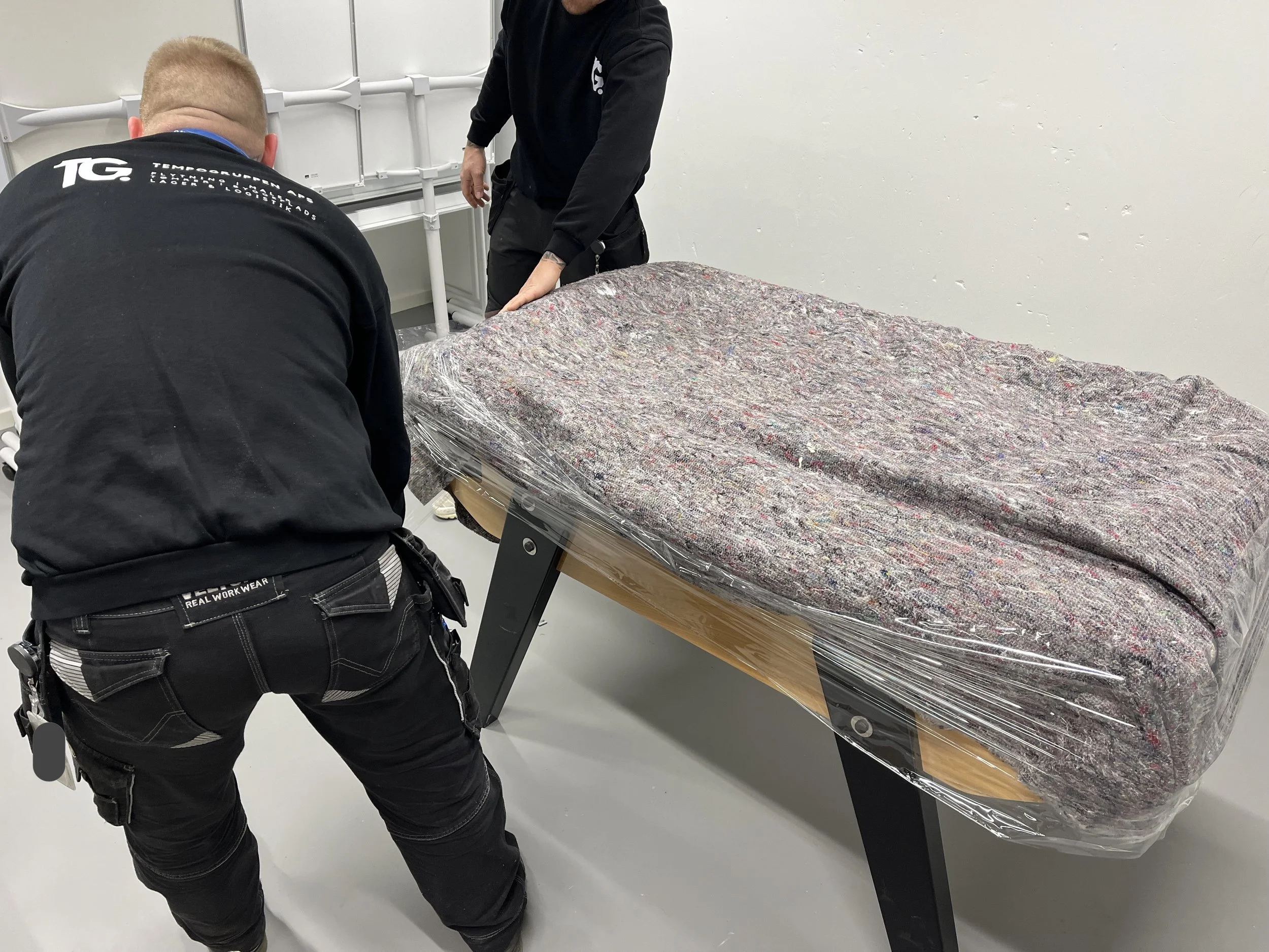 To personer pakker et bolig- eller kontorunderlag ind i plastik, mens de arbejder sammen i et rum med hvide vægge.