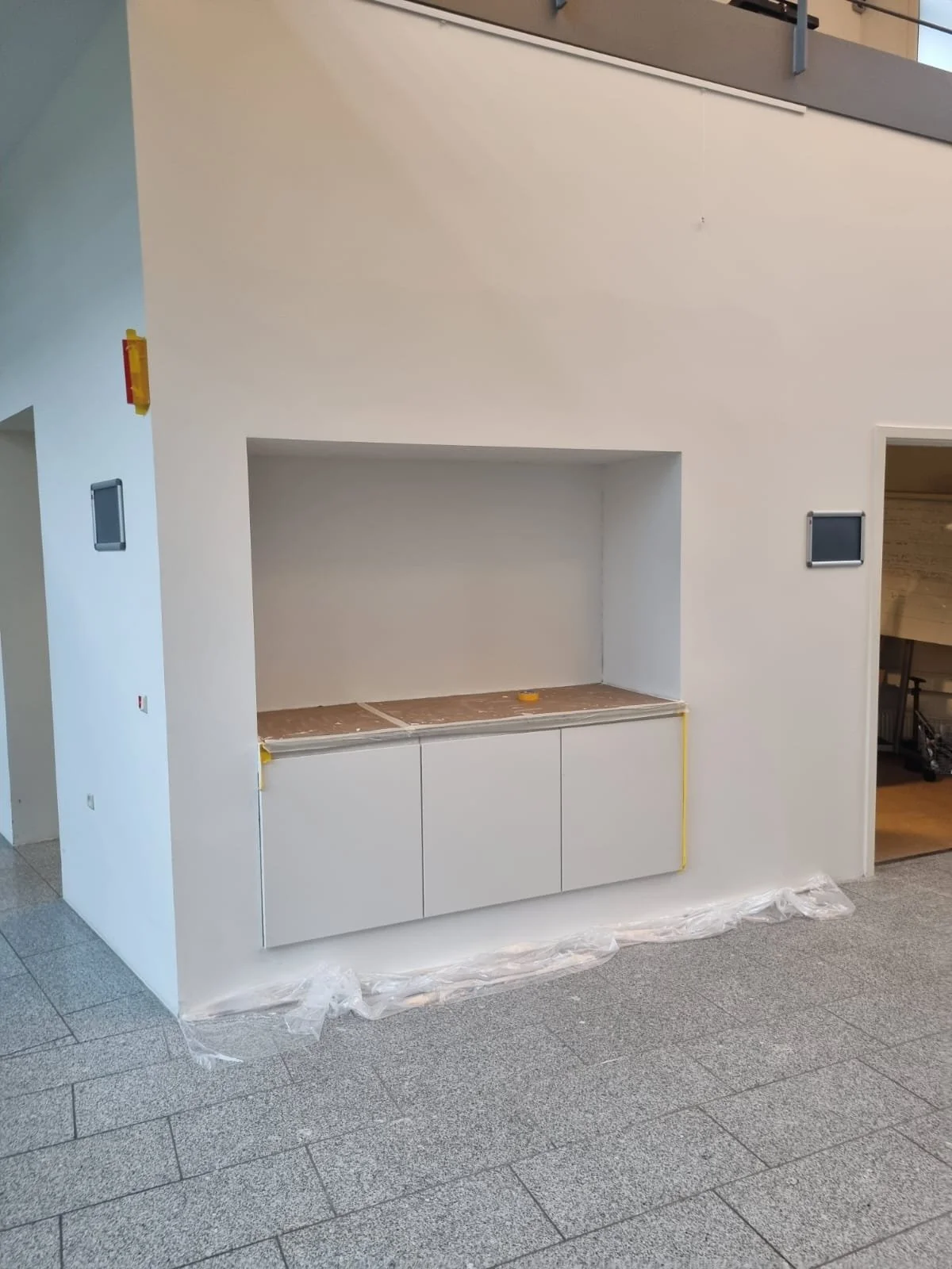 Ufærdig sammenbygning af et hvidt skab under en indbygget hylde, med plastfolie på gulvet foran.