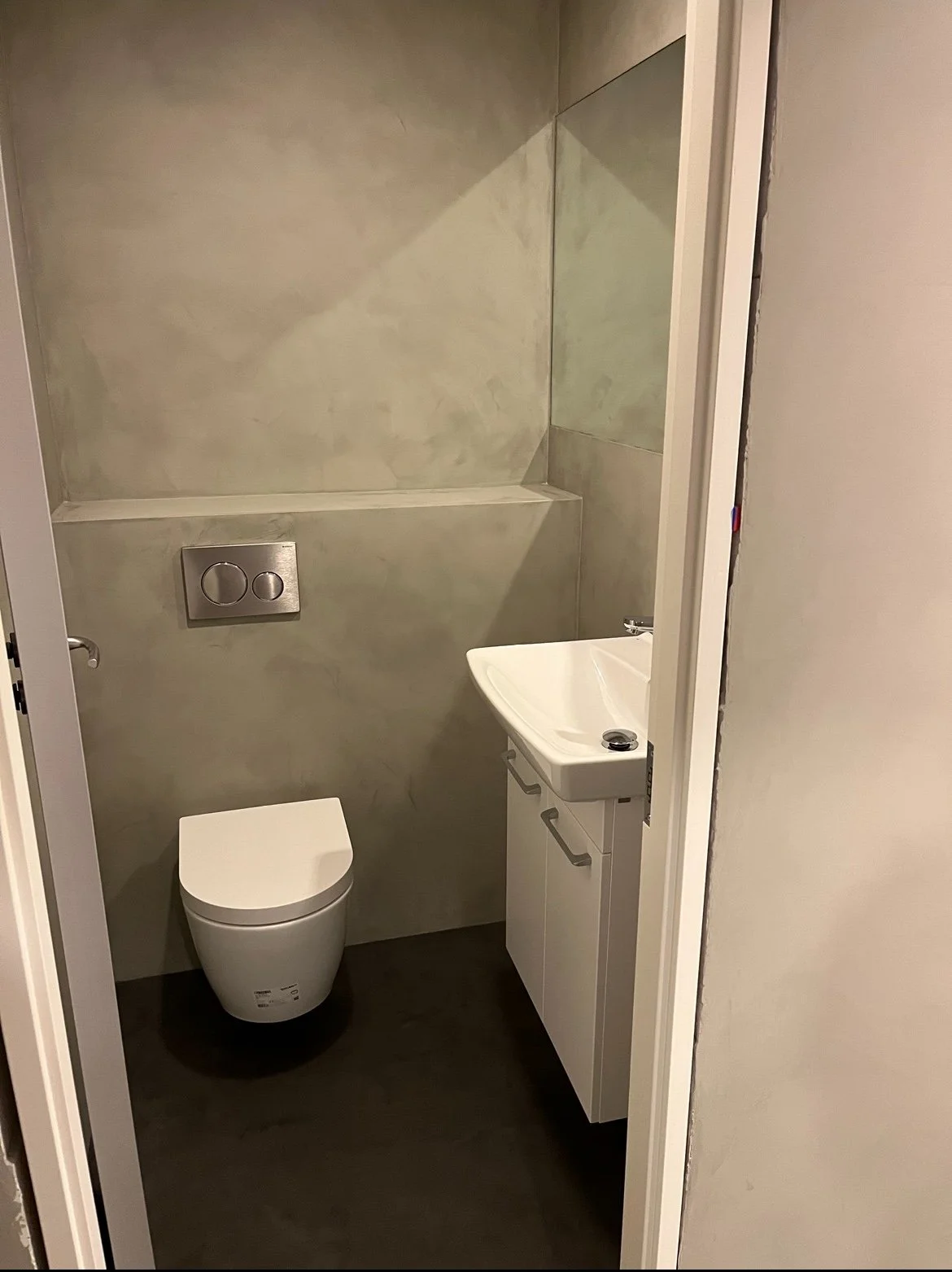 En lille toilet med vægtoilet, vask og spejl, med lyse vægge og mørkt gulv.