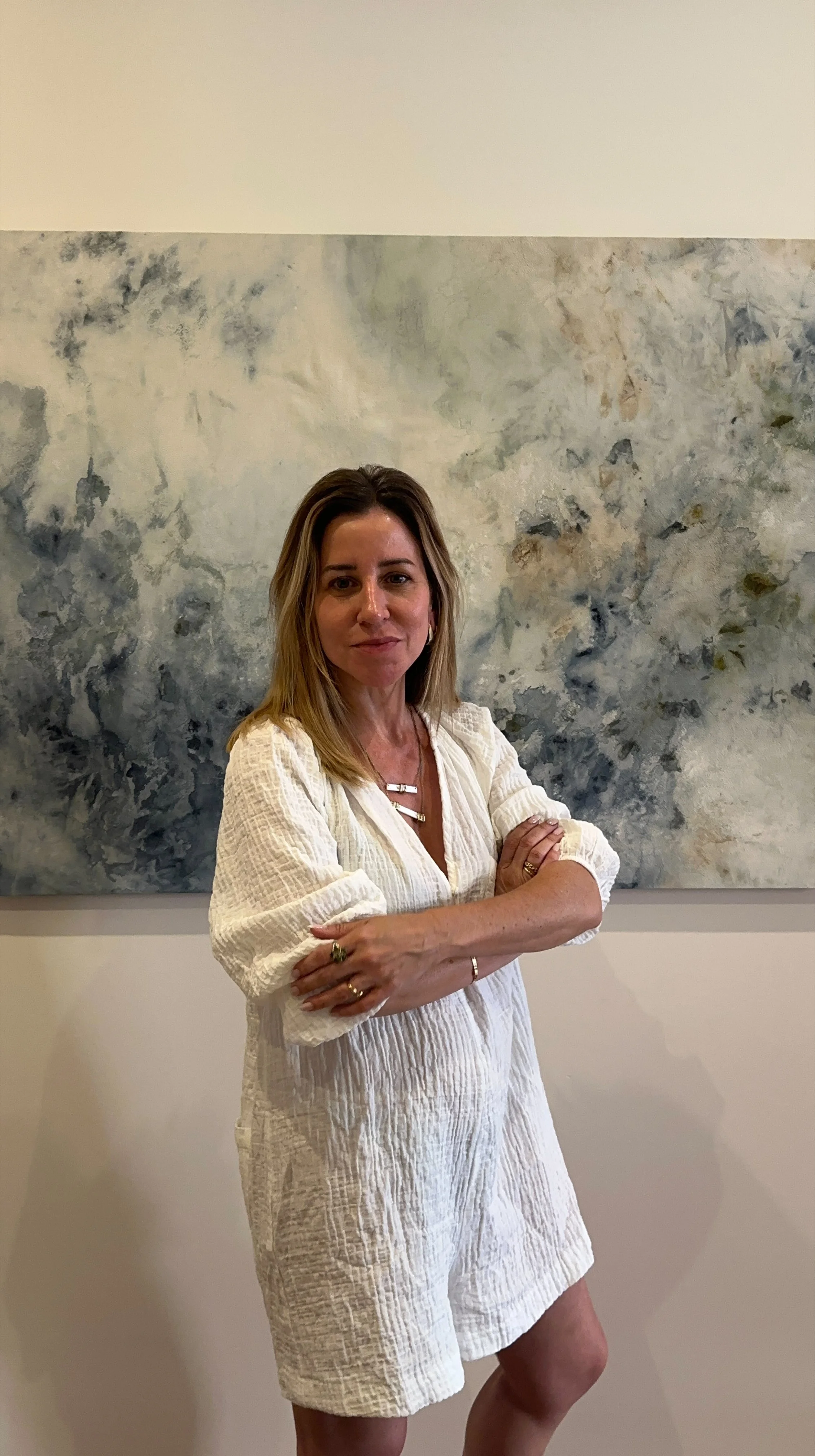 Mujer de cabello rubio y peinado suelto, vestida con un conjunto blanco, posa con los brazos cruzados frente a un fondo de piedra y una pared blanca.