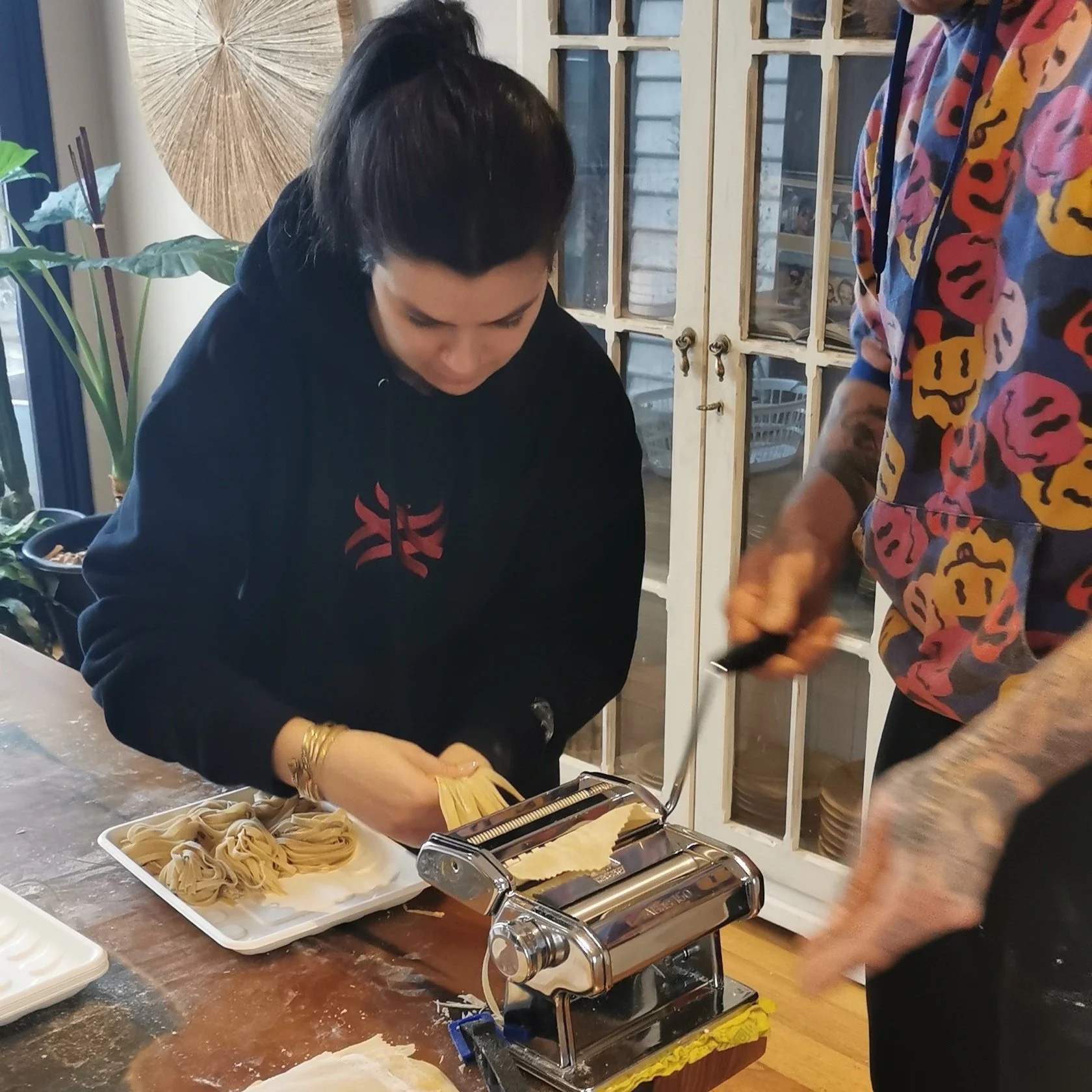 A woman preparing fresh pasta using a pasta machine while a man assists, in a kitchen with natural light, plants, and a glass cabinet.