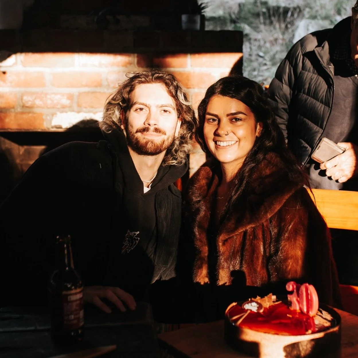 A man and woman sitting at a table in a cozy, dimly lit restaurant or bar with a brick wall in the background. They are smiling and close together, with birthday decorations and a red cake in front of them.