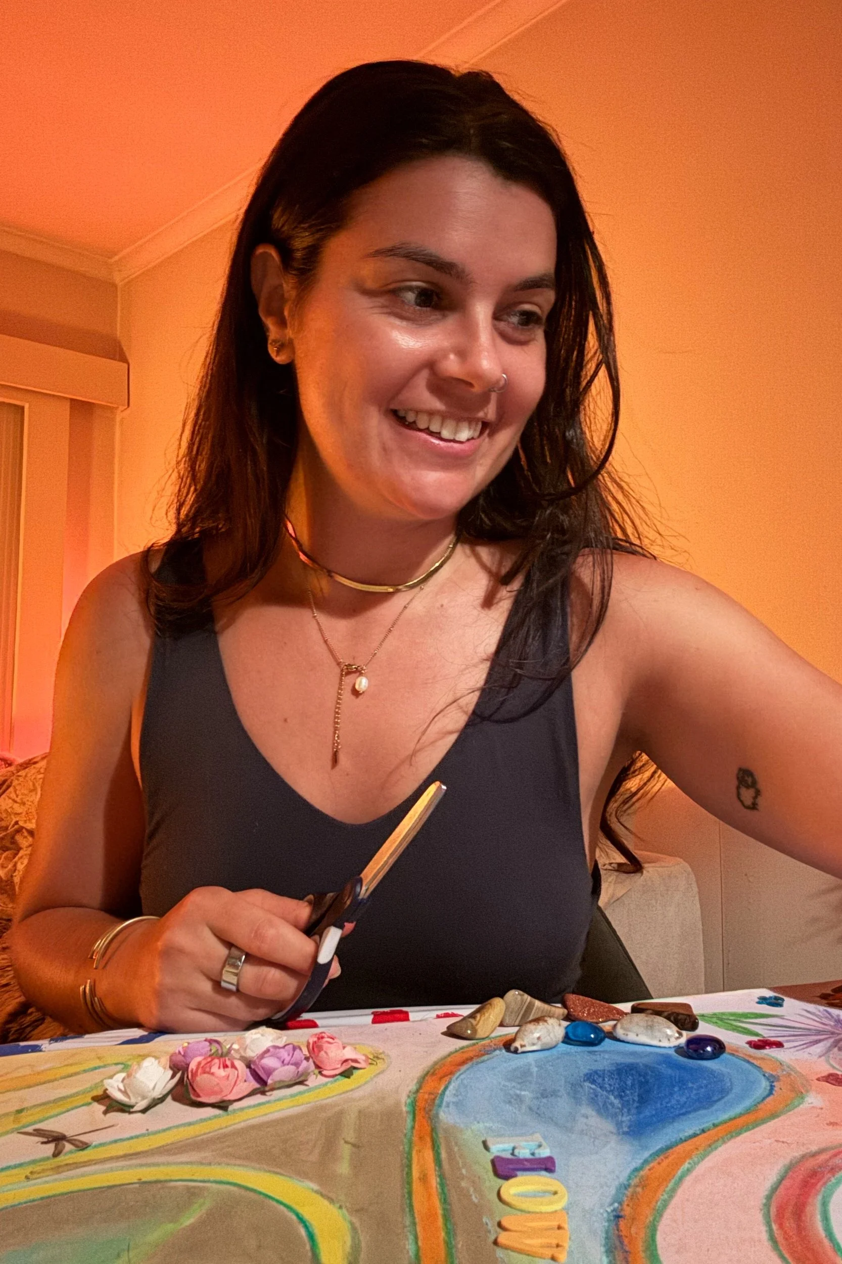 Woman with dark hair, wearing a black top and layered necklaces, sitting at a table with painted designs and various small objects, smiling and looking down, in a warmly lit room.
