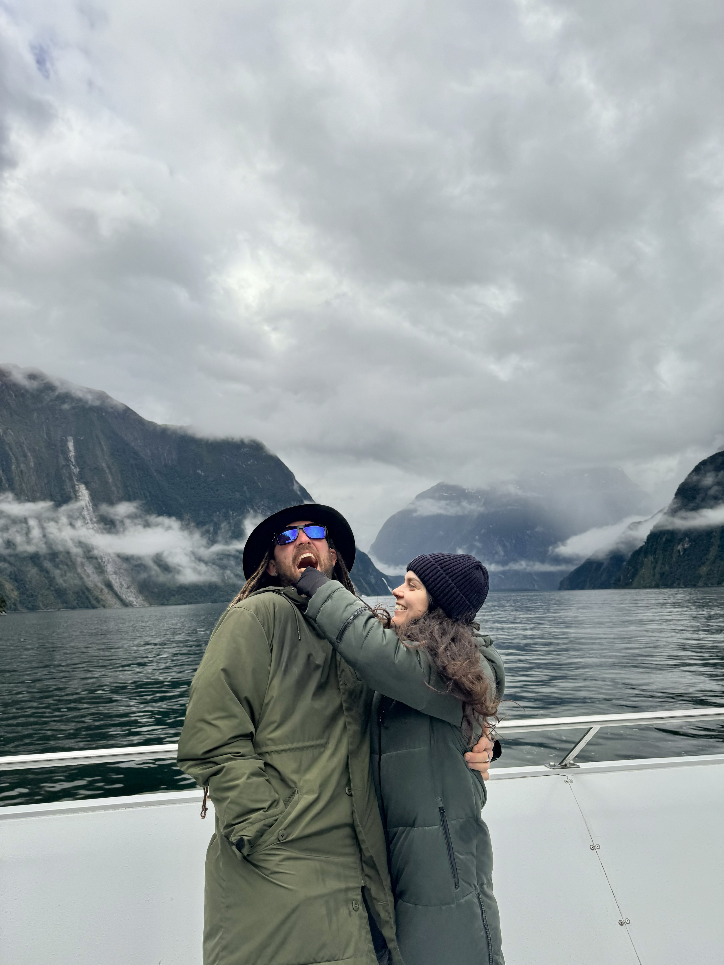 A man and woman are on a boat in a scenic fjord, with misty mountains and cloudy sky in the background. The woman is grabbing the man's chin in a playful manner, and they are both smiling.