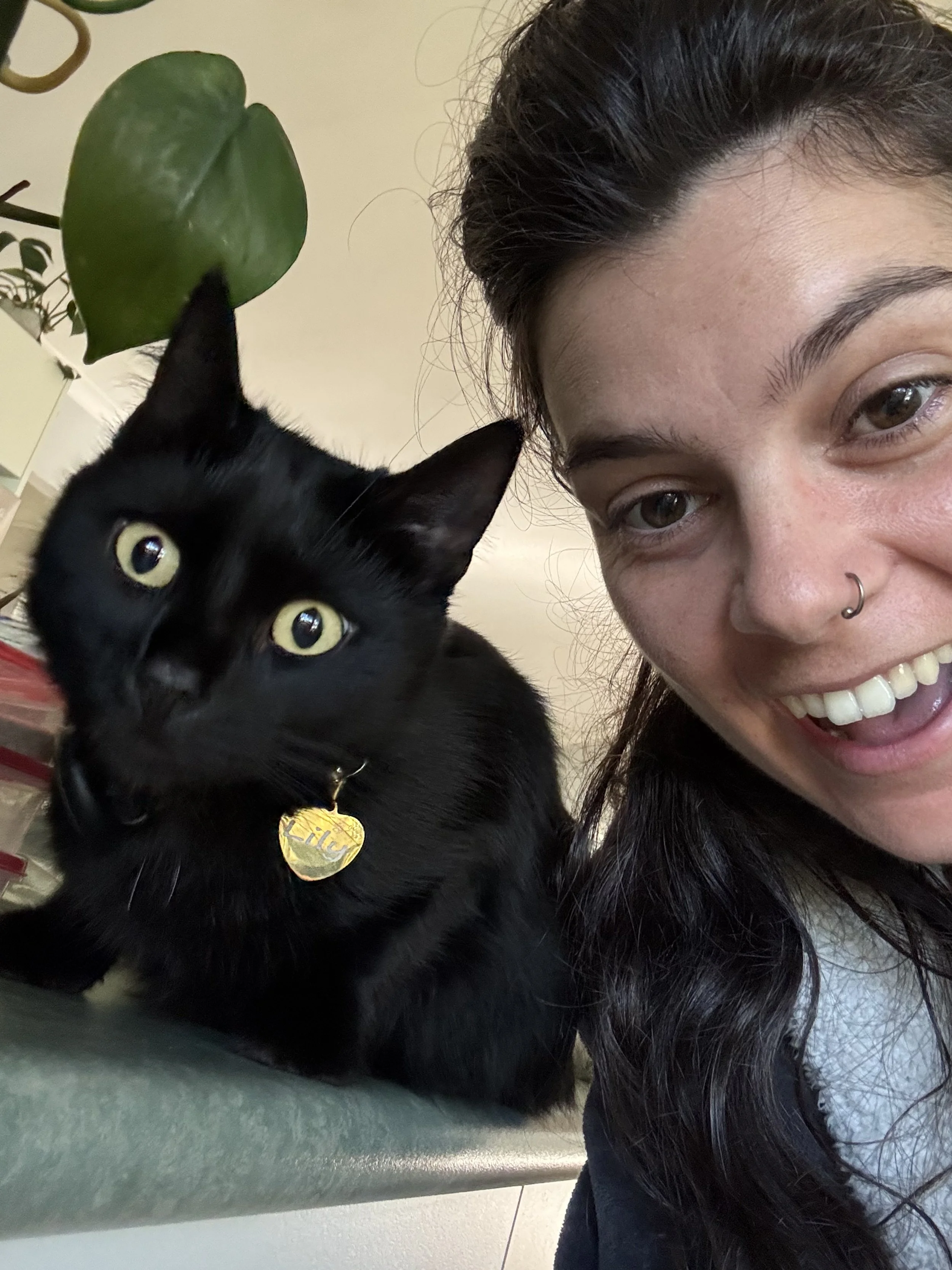 A woman with a nose ring smiling next to a black cat with yellow eyes, wearing a gold heart-shaped collar tag, sitting on a surface with a houseplant in the background.