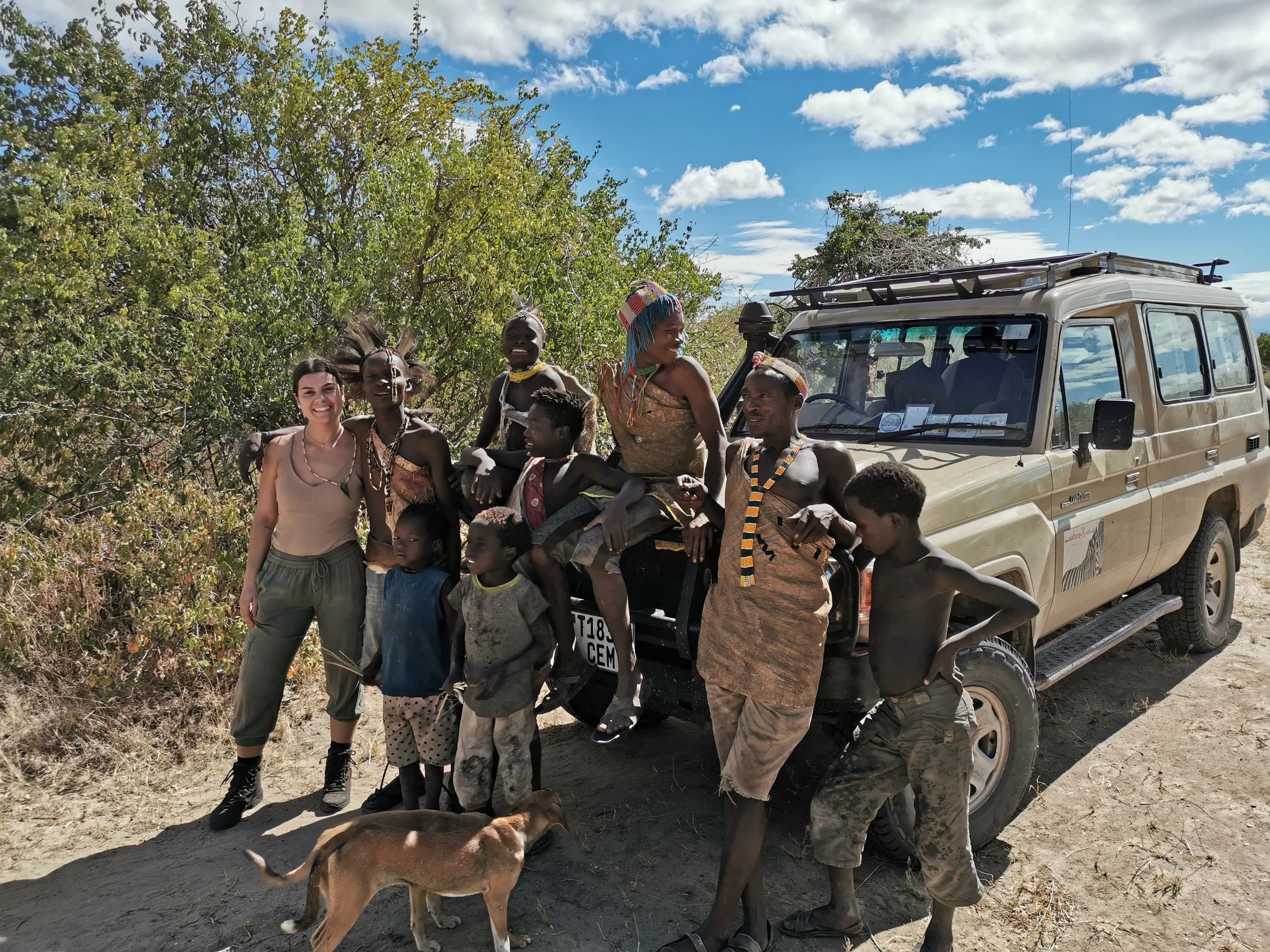 Group of people, including children, standing next to a beige off-road vehicle on a dirt road with trees and a partly cloudy sky in the background.