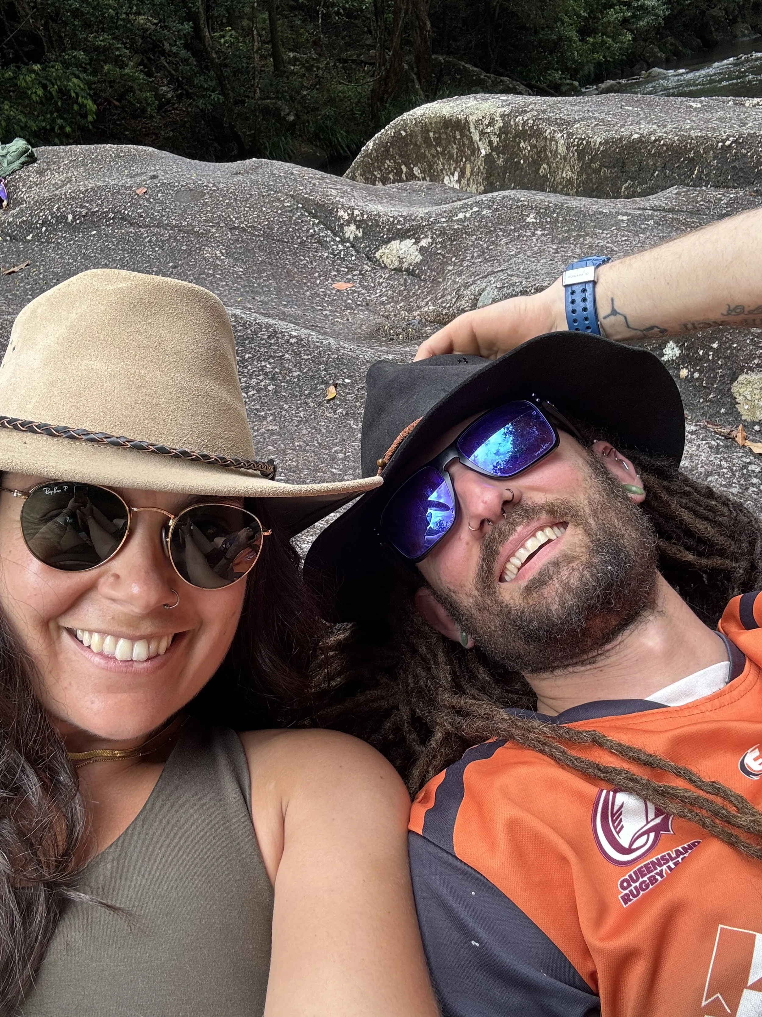 Two smiling people, a woman and a man, taking a selfie outdoors on a rocky surface. The woman is wearing a beige wide-brimmed hat and sunglasses, and the man is wearing a black hat, sunglasses, and an orange sports jersey. The background features lar