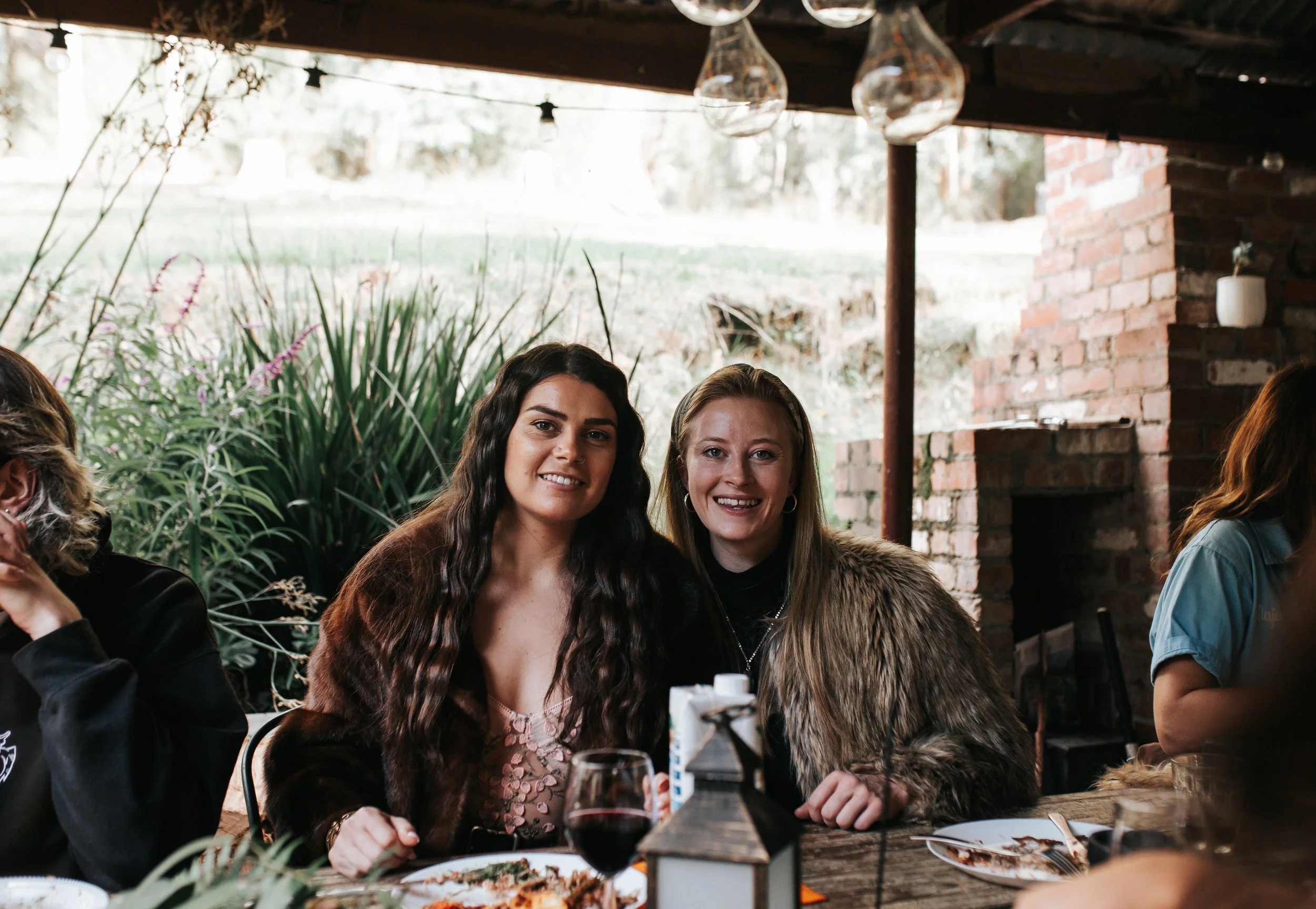 Two women sitting at a table during a meal, smiling for the camera outdoors with greenery and brick structure in the background.