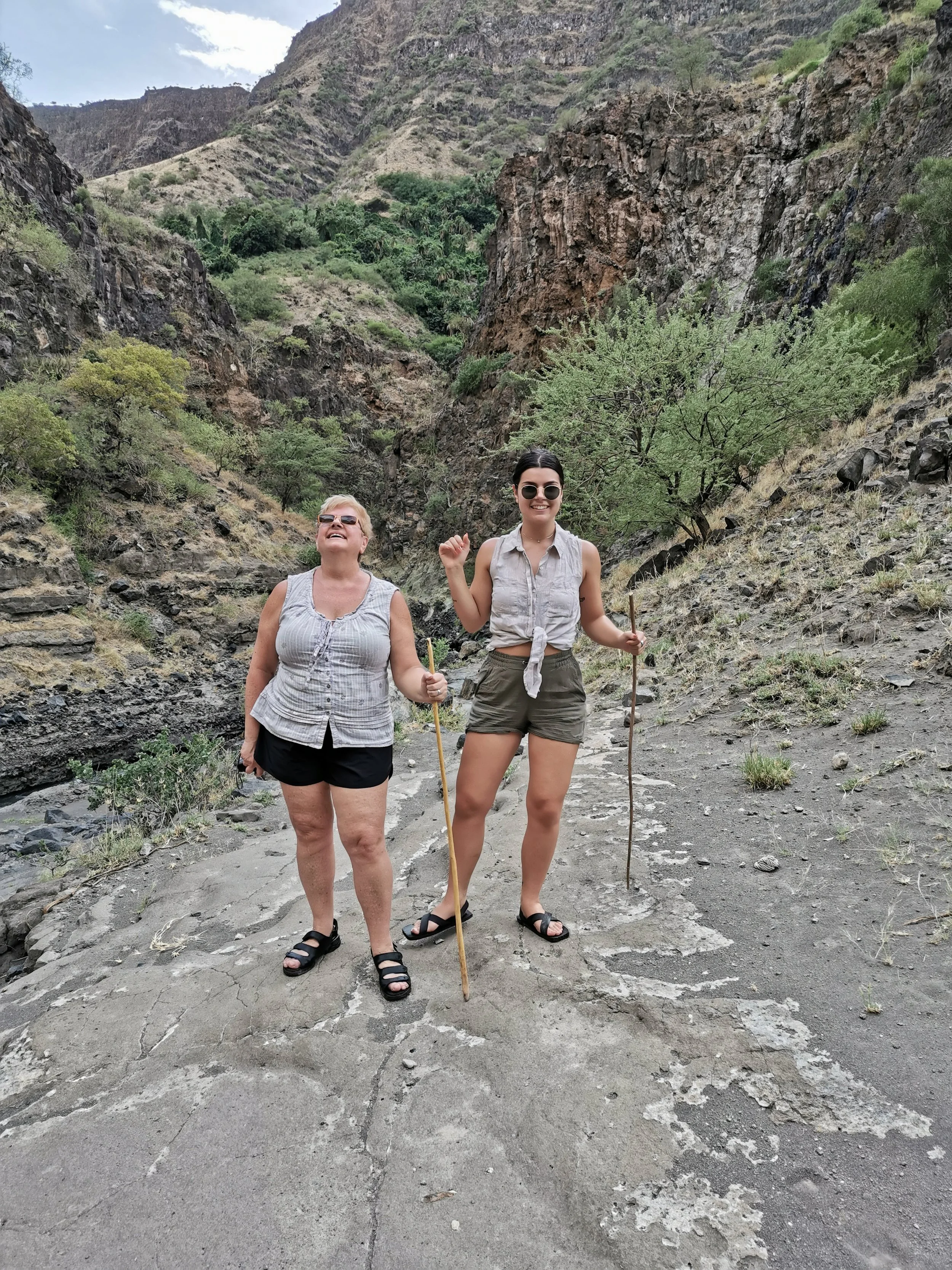 Two women hiking in a canyon with rocky cliffs, sparse vegetation, and dry terrain, smiling and holding hiking sticks.