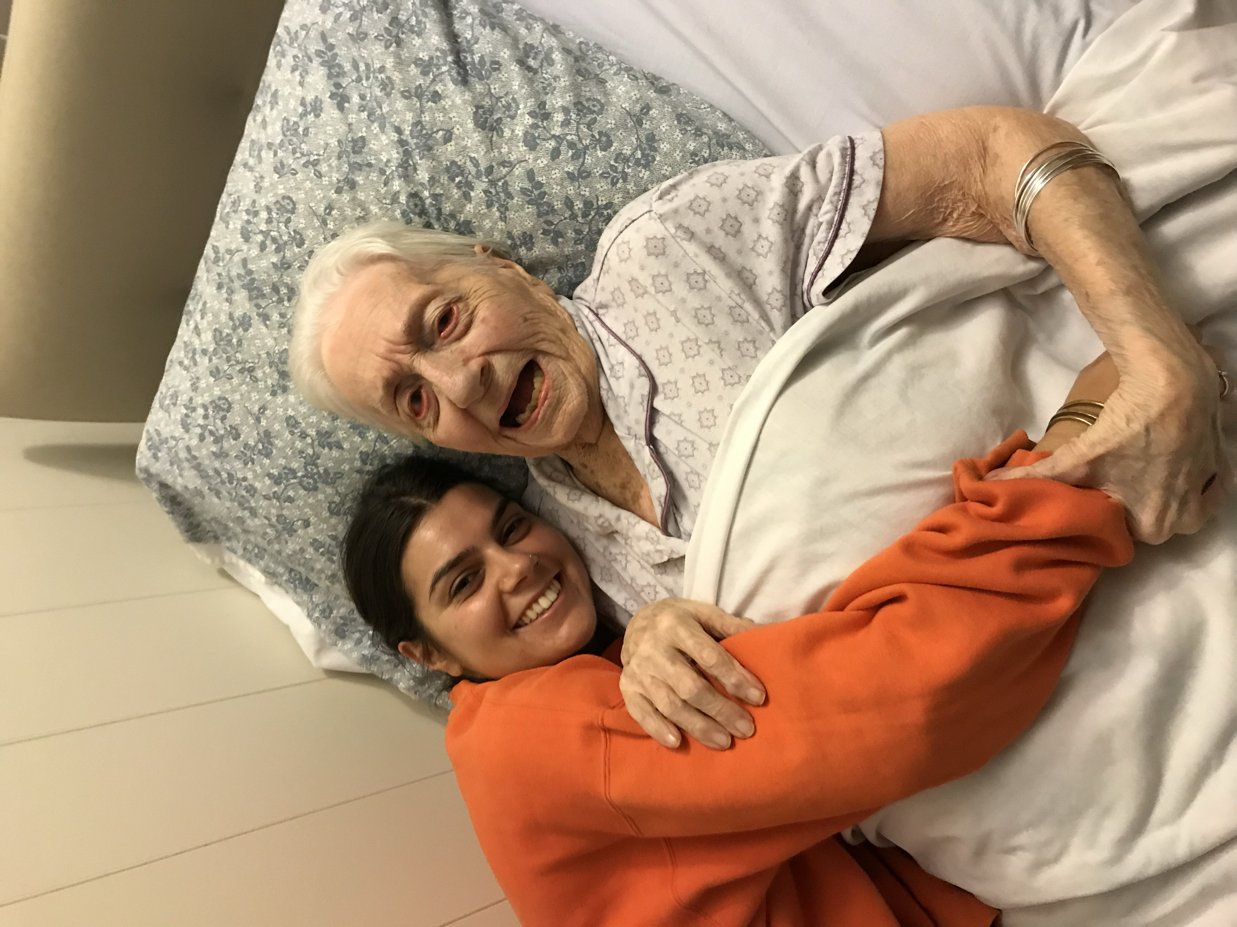A young woman and an elderly woman are lying in bed together, smiling and embracing. The elderly woman is on a hospital bed with a patterned pillow and a blanket, and the young woman is beside her, holding her hand.