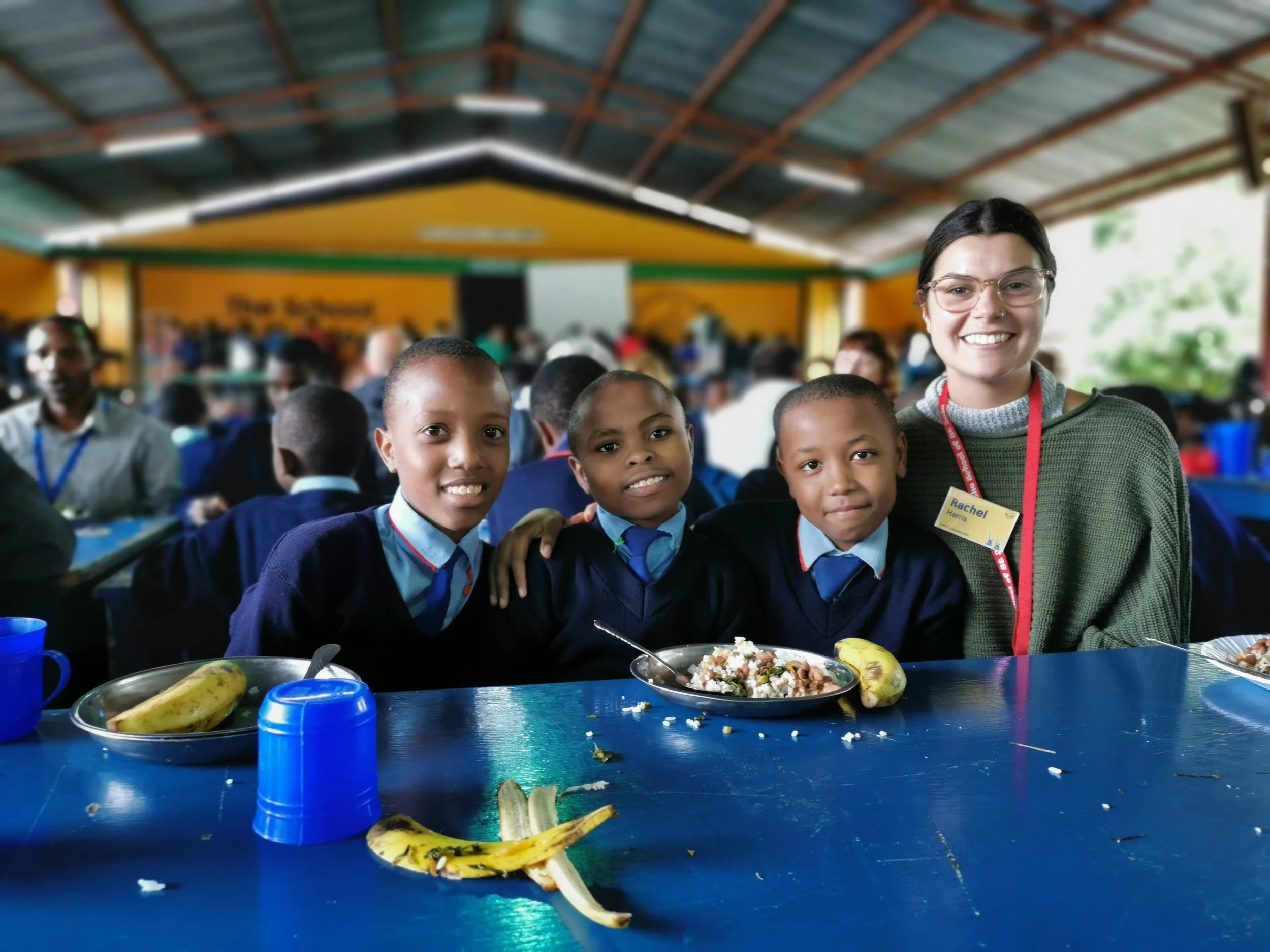 A group of four people, three school children in uniform and a woman, smiling at the camera at a school cafeteria. They are sitting at a blue table with bananas and a tray of food in front of them, with many students in the background.