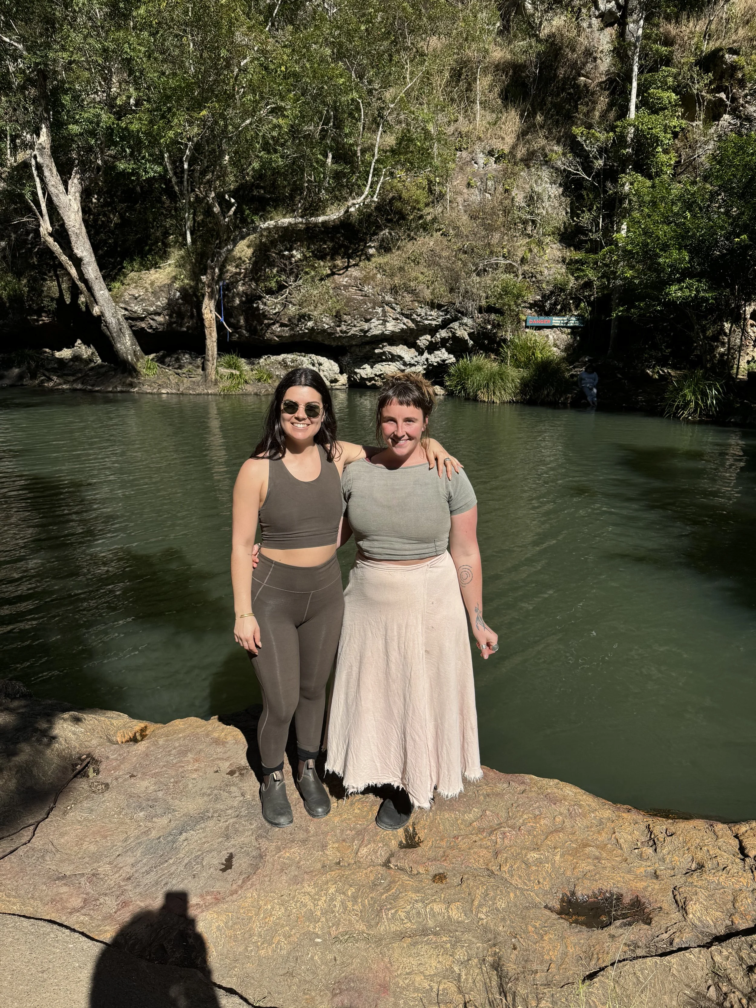 Two women standing on a rocky riverbank, smiling at the camera, arm around each other, in a natural outdoor setting with green trees and water behind them.