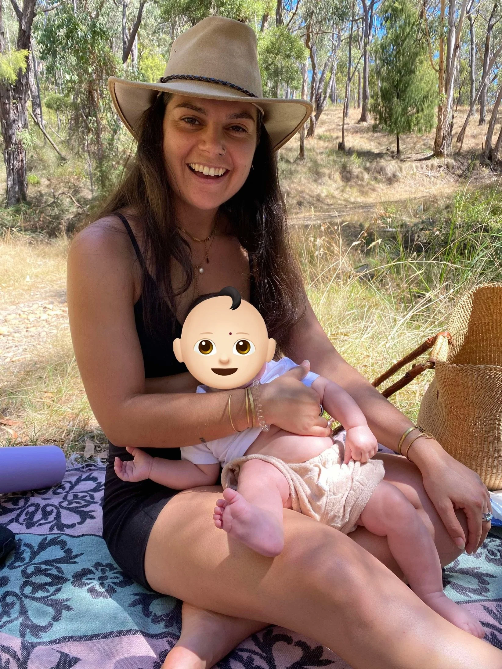 A woman in a wide-brimmed hat smiling while holding a baby with a cartoon face sticker outdoors in a wooded area, sitting on a blanket with a wicker chair nearby.
