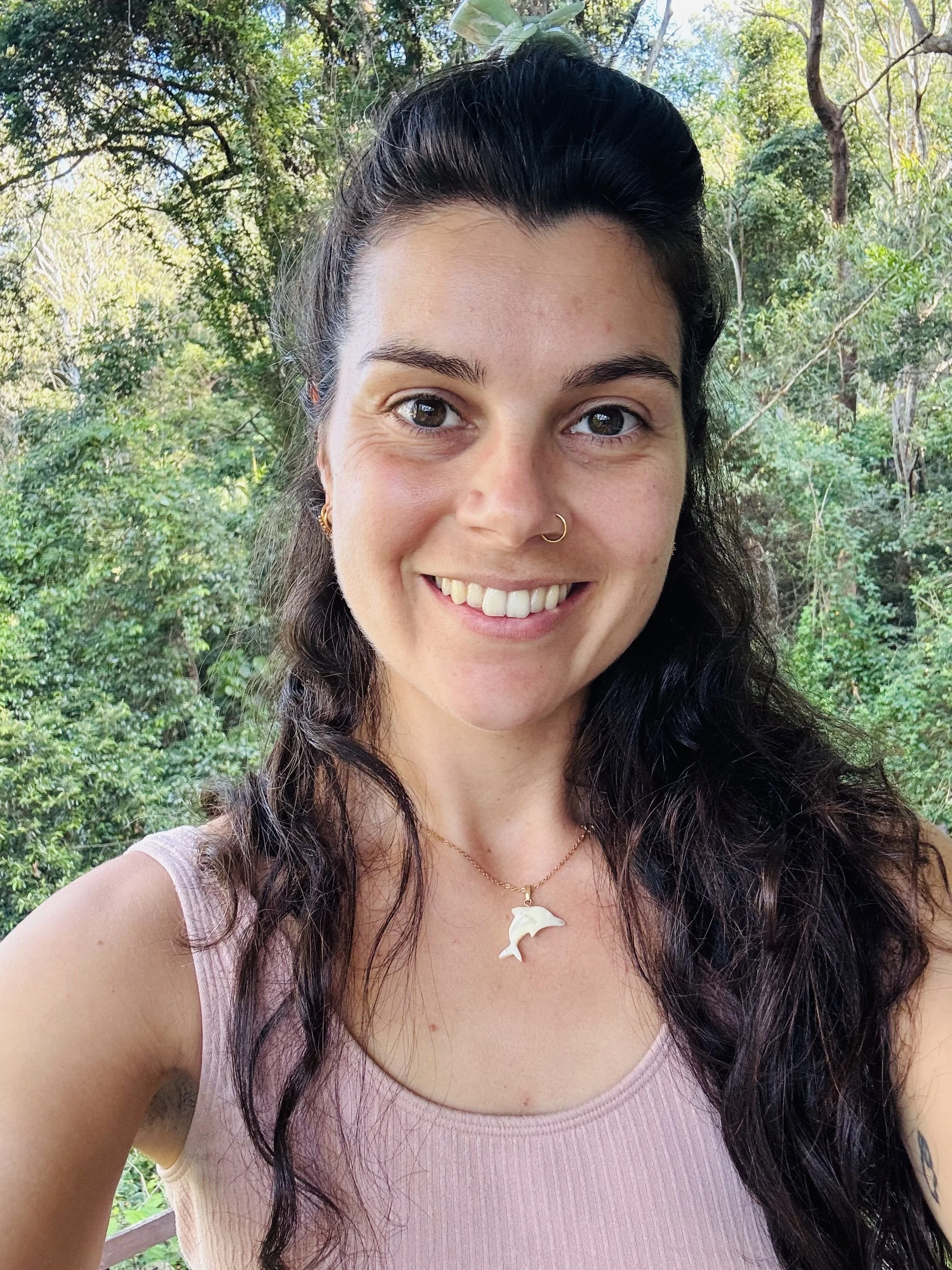 A woman smiling outdoors with a background of trees and greenery, wearing a necklace with a dolphin pendant.