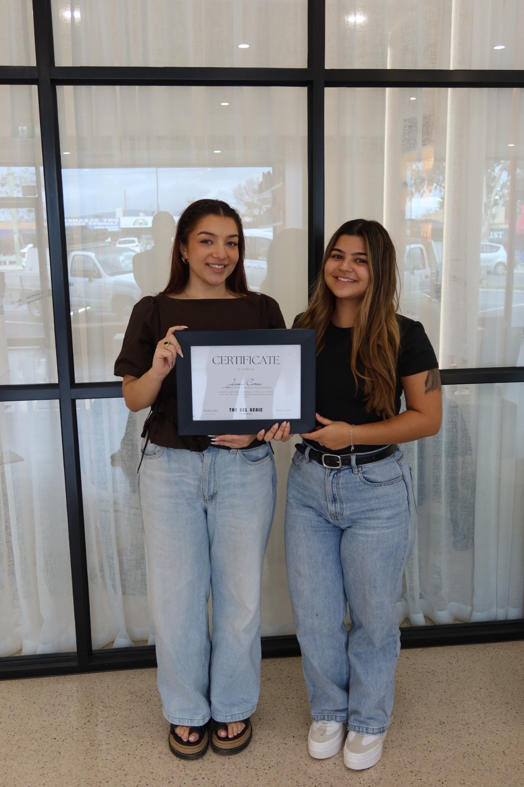 Two women stand side by side in front of a window with curtains, holding a certificate of achievement. The woman on the left has long brown hair, wearing a black top and light jeans. The woman on the right has long wavy brown hair, wearing a black shirt and light jeans with sneakers. They are smiling for the camera.