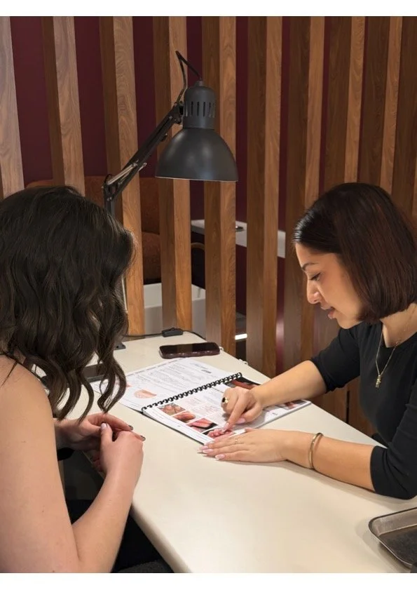 A makeup artist demonstrating makeup techniques to a woman seated at a table in a salon with vertical wooden slats in the background and a desk lamp overhead.