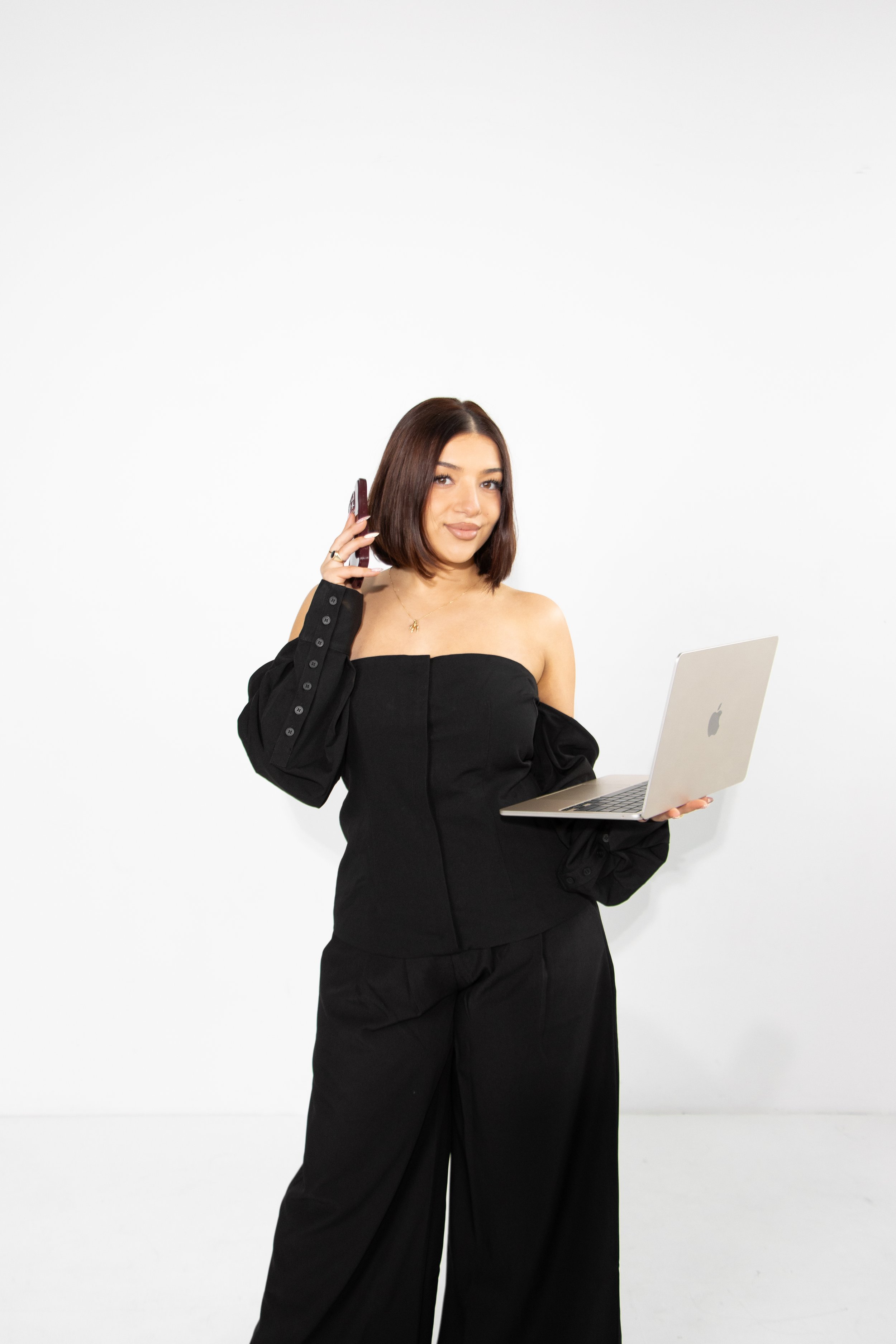 A woman with shoulder-length brown hair, wearing an off-shoulder black outfit and wide-legged black pants, stands against a plain white background. She holds a silver laptop in her left hand and a phone to her ear with her right hand, smiling slightly.