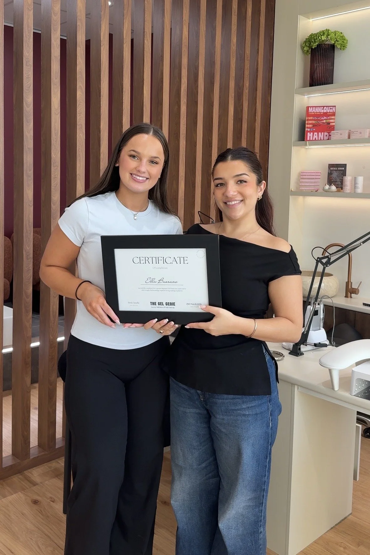 Two women smiling and holding a certificate indoors, one in a light blue shirt and the other in a black shirt, with a wooden wall and shelves in the background.