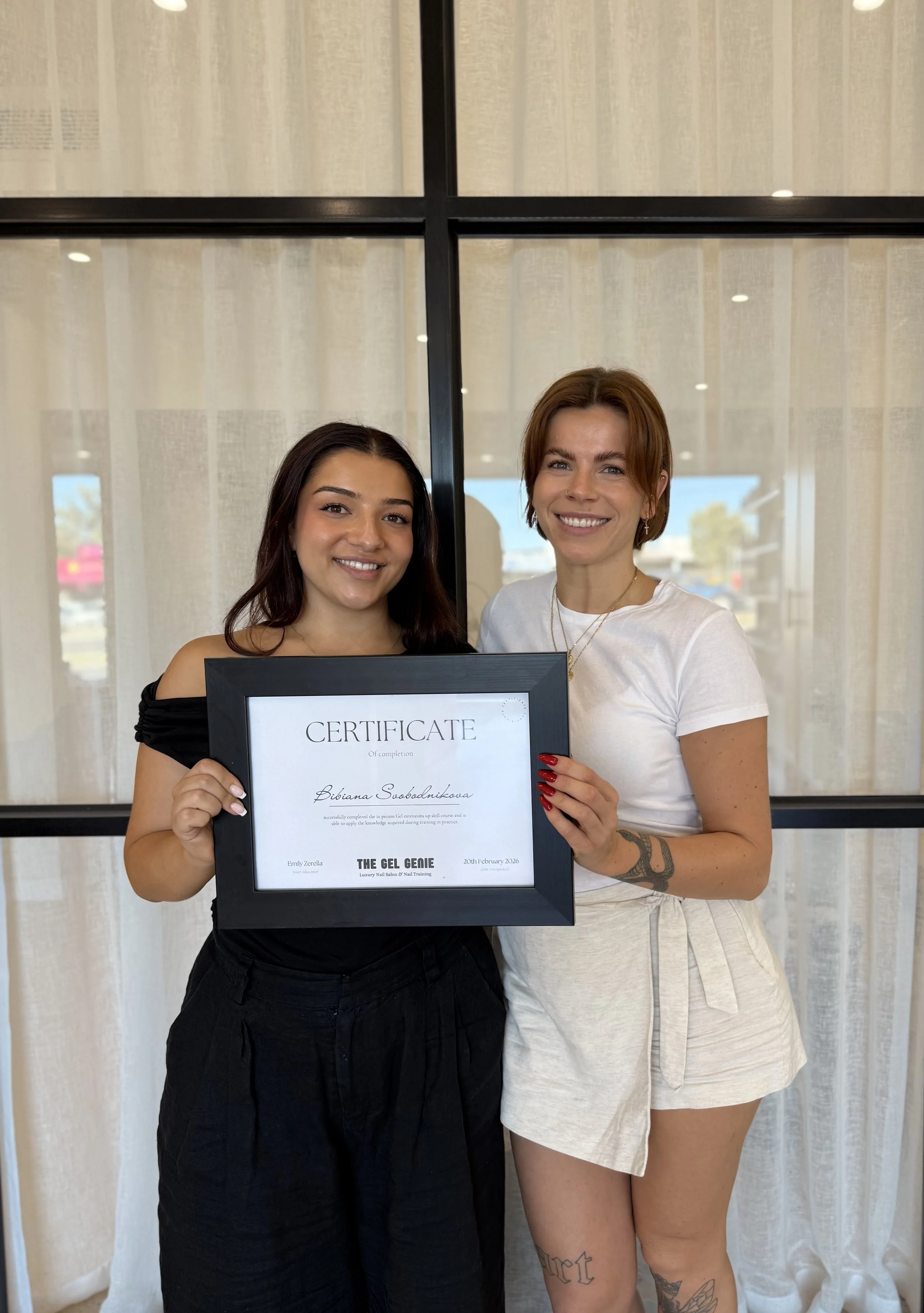 Two women smiling and holding a framed certificate in front of white curtains. One woman has long dark hair and is wearing a black top, the other has short red hair and is wearing a white T-shirt with tattoos on her leg.